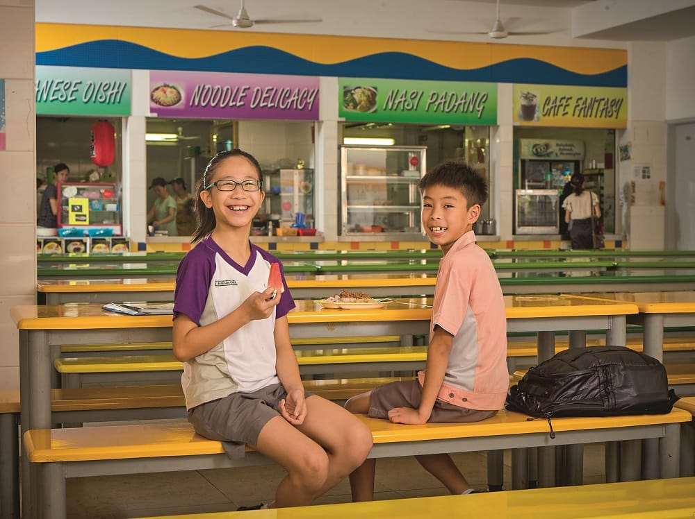 Two smiling children sit on yellow benches in a school canteen with food stalls including Noodle Delicacy and Nasi Padang.