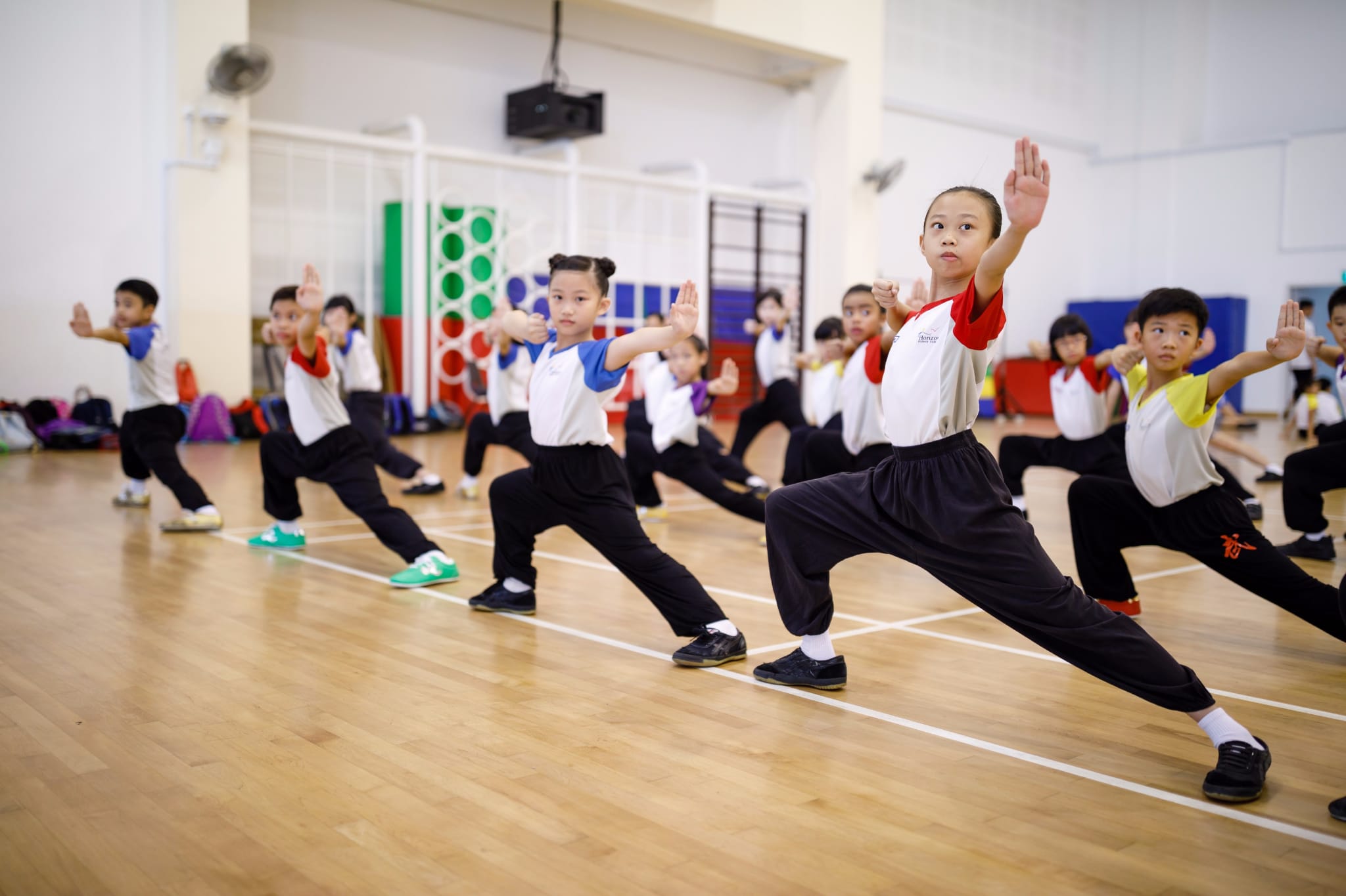 Children in white and black uniforms practice martial arts in a wide stance with raised hands in a school gymnasium.