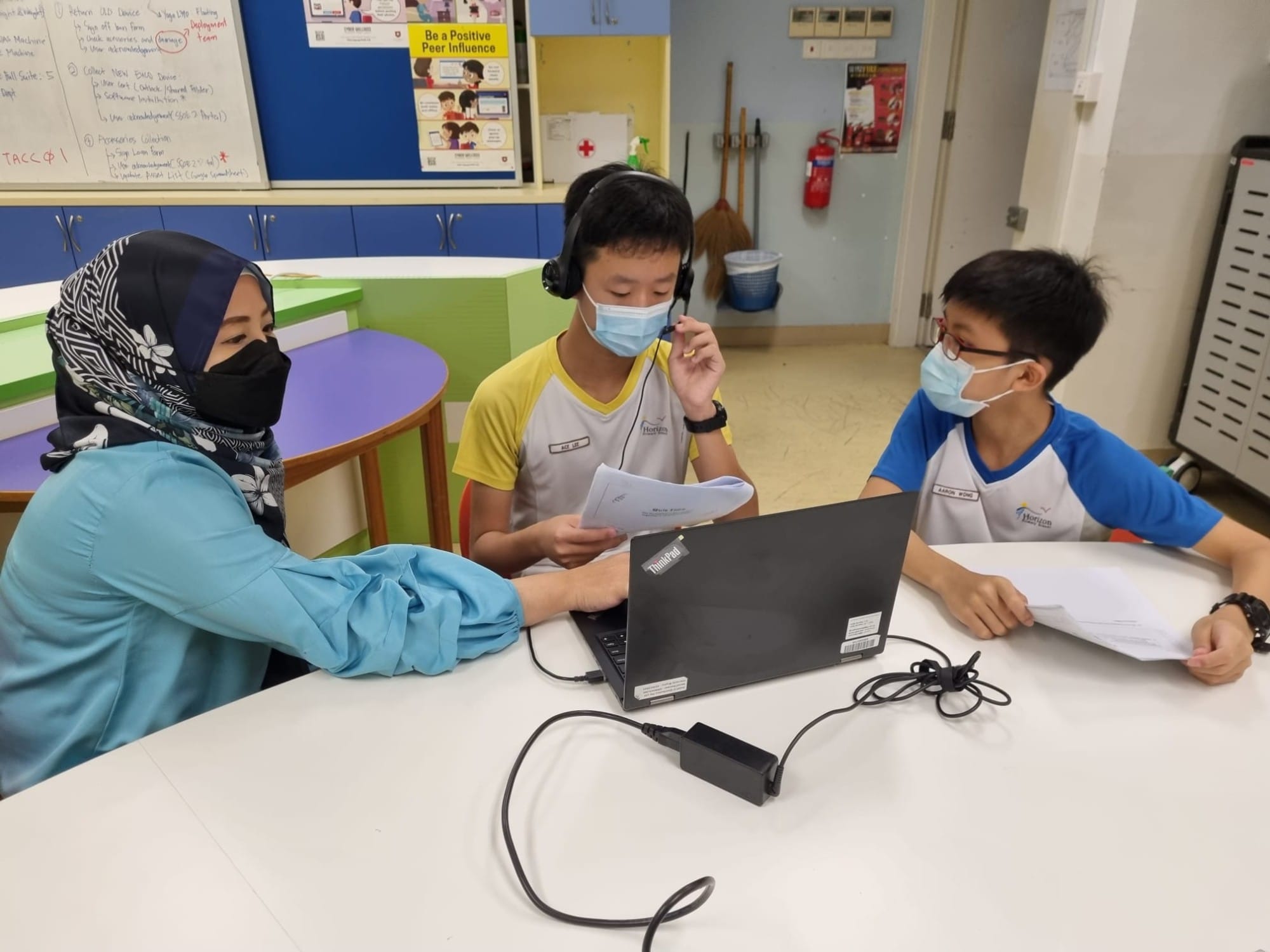 Three masked students sit at a table with a ThinkPad laptop, one wearing a headset and holding papers in a classroom.