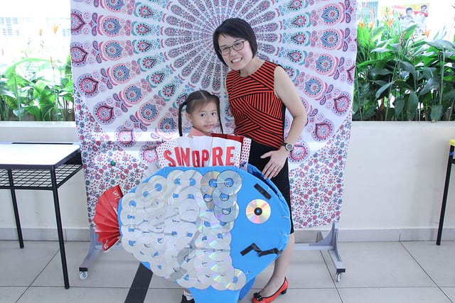 Woman and young girl posing with a blue cardboard fish decorated with CDs in front of a floral backdrop.
