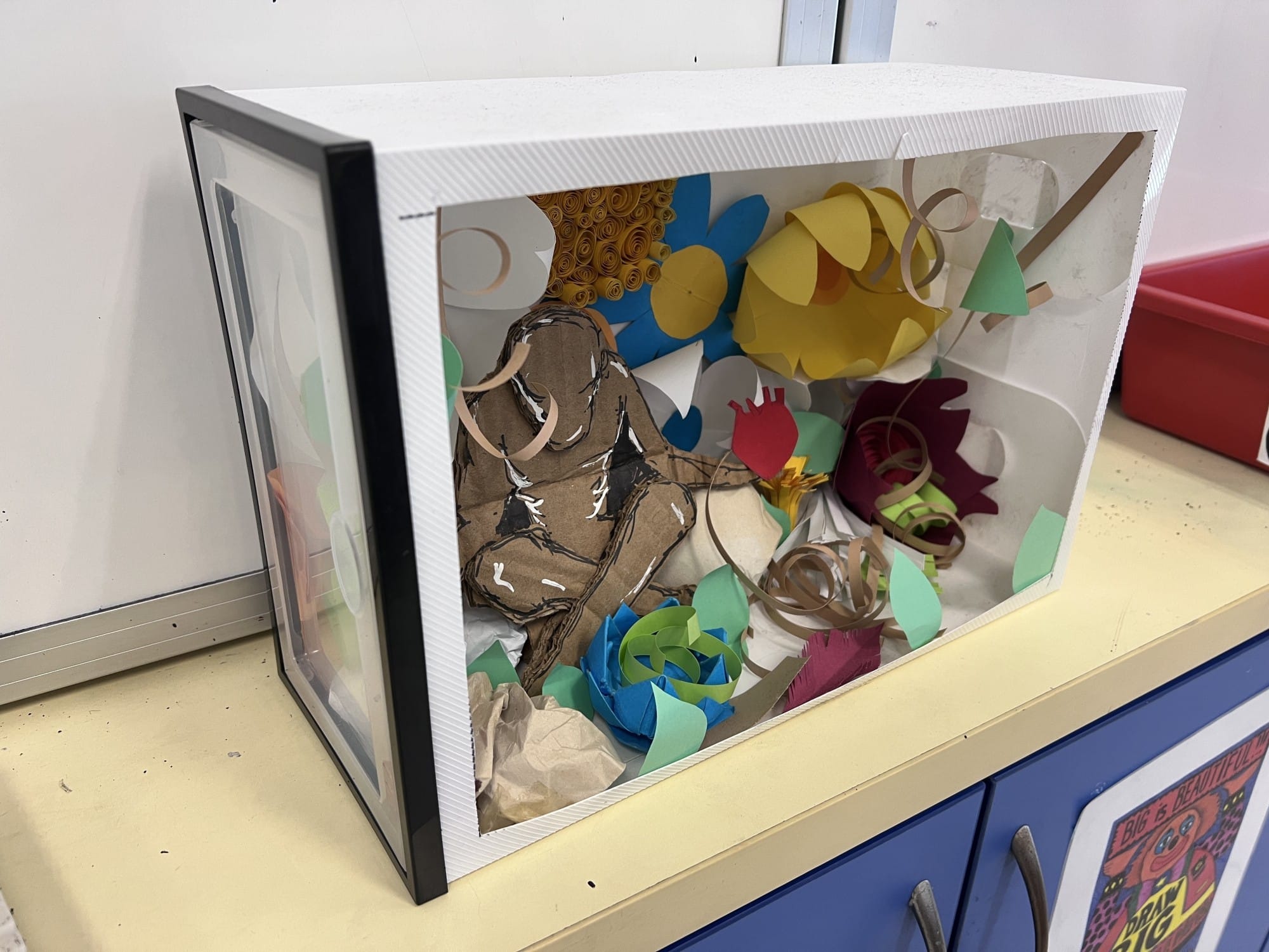 A white box diorama with a clear front holds colorful paper flowers, curled strips, and a cardboard human figure cutout.