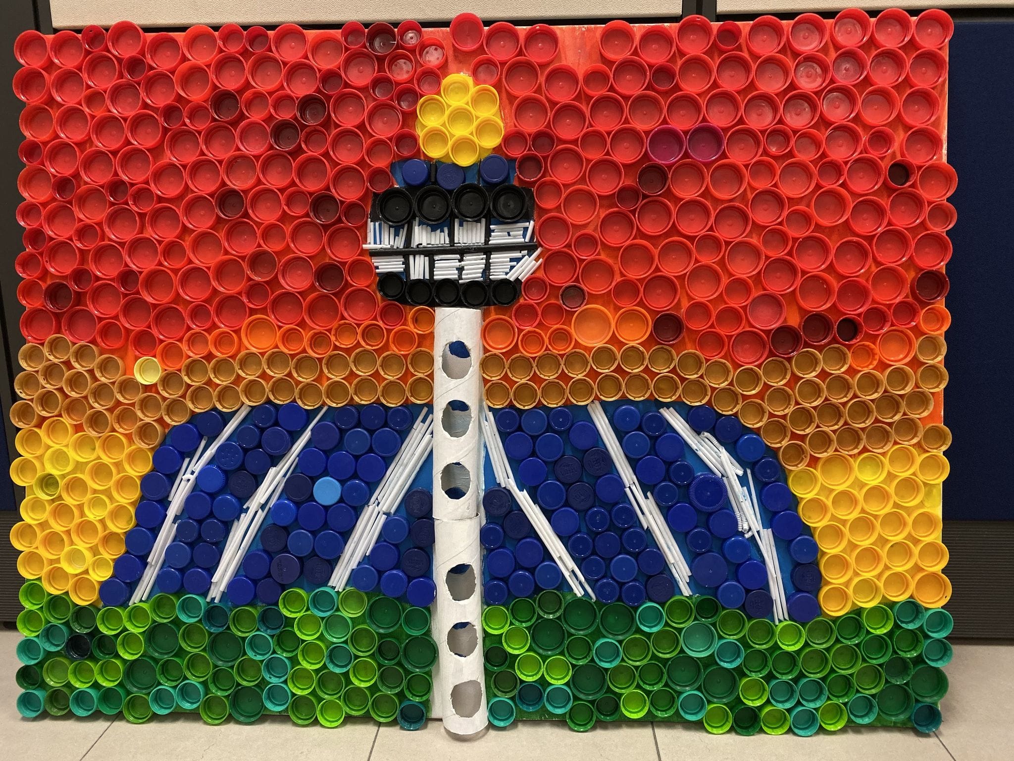 Mosaic artwork of a tower with rainbow-colored rows of plastic bottle caps arranged in red, orange, yellow, blue, and green.