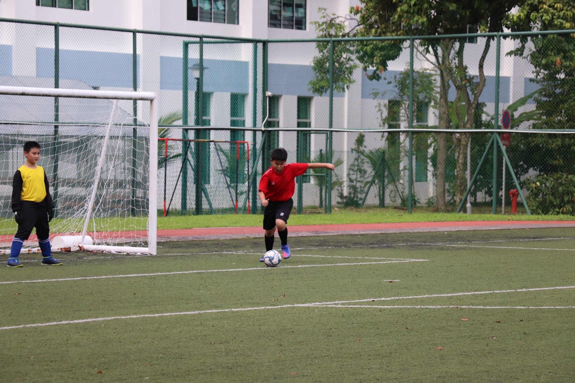 Boy in red shirt kicking a soccer ball on a green turf field near a white goal, with a goalkeeper in yellow watching.