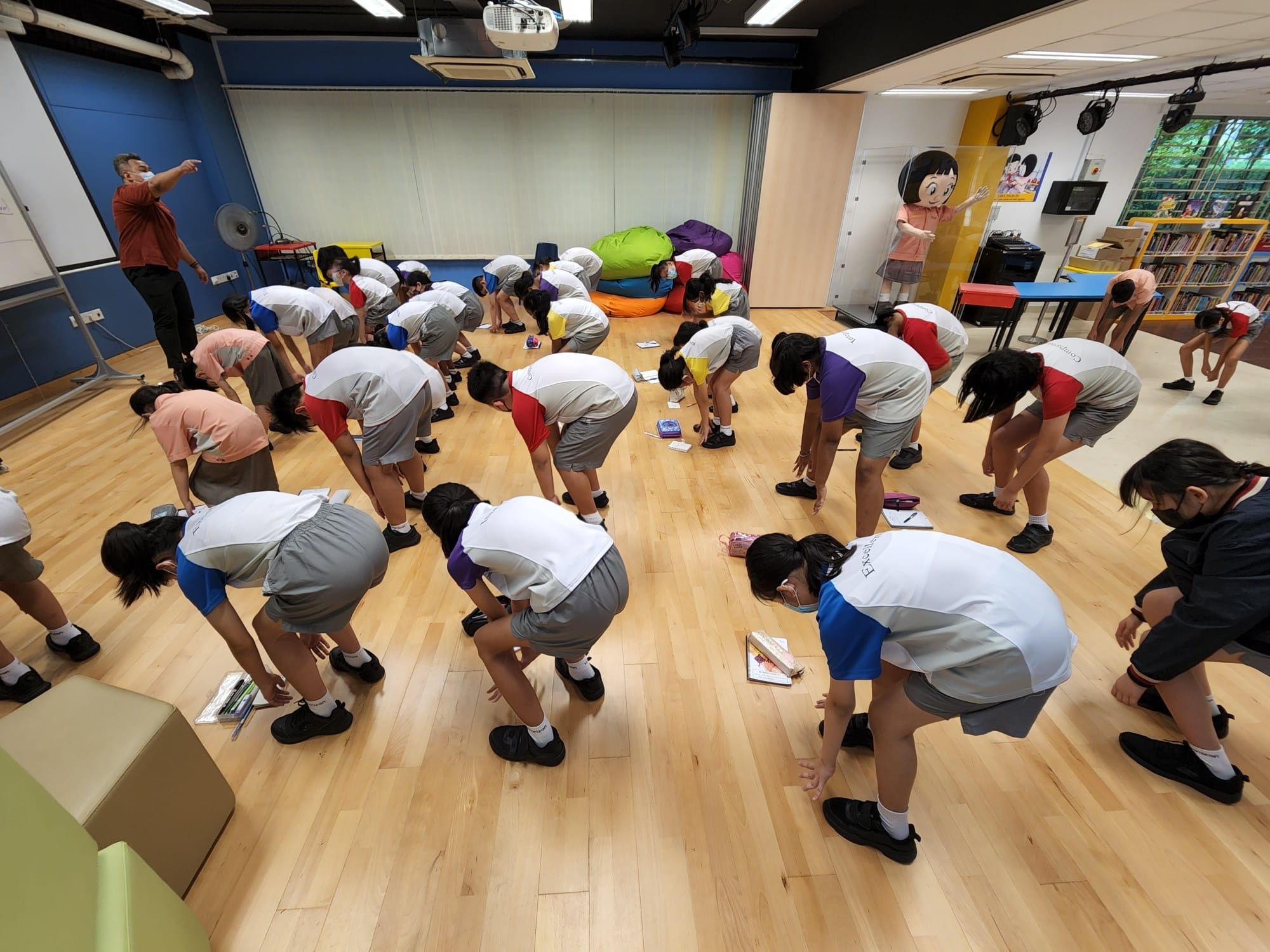 A group of school children in uniforms bow forward on a wooden floor while a teacher points directions in a classroom.
