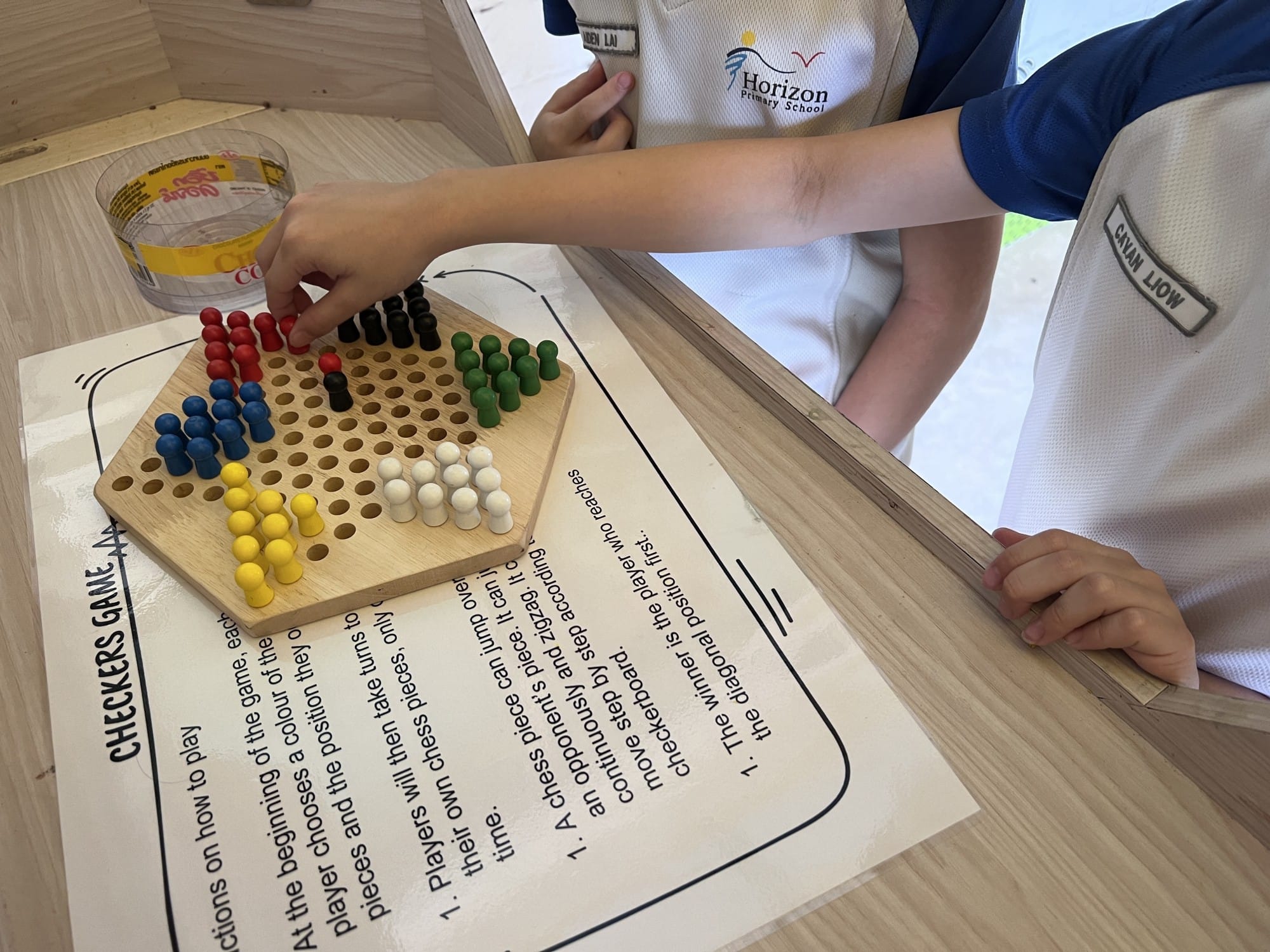 Two children in Horizon Primary School uniforms playing Chinese checkers on a wooden board with colorful pegs.