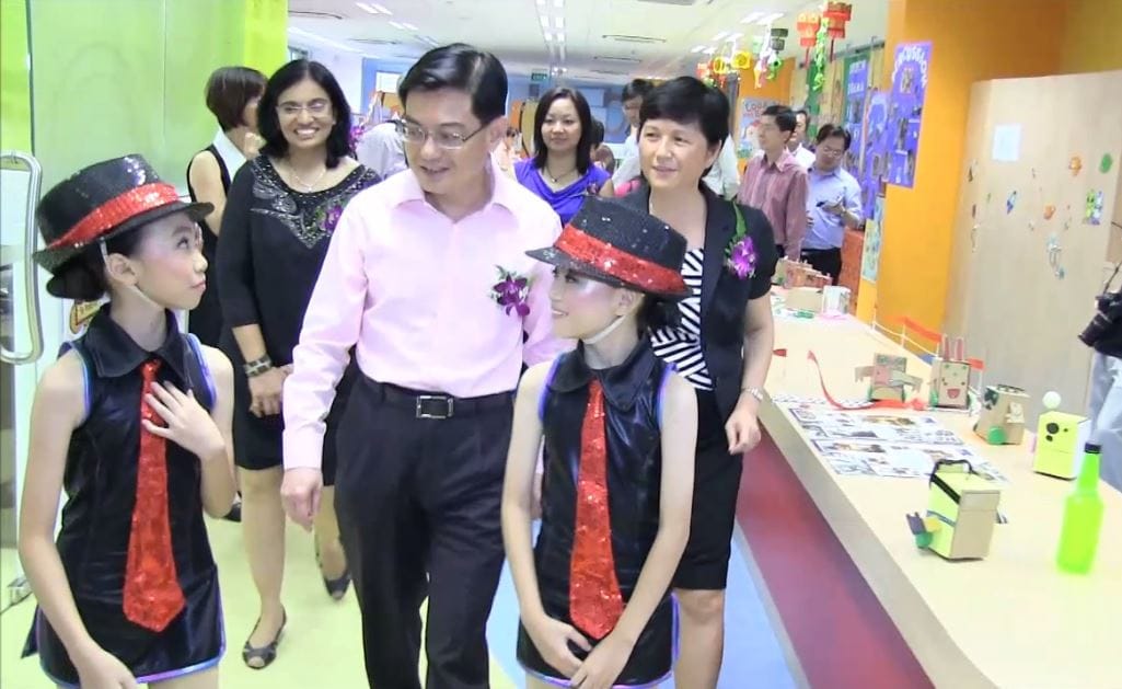 Adults and two girls in black outfits with red sequin ties and hats walk through a colorful school hallway.