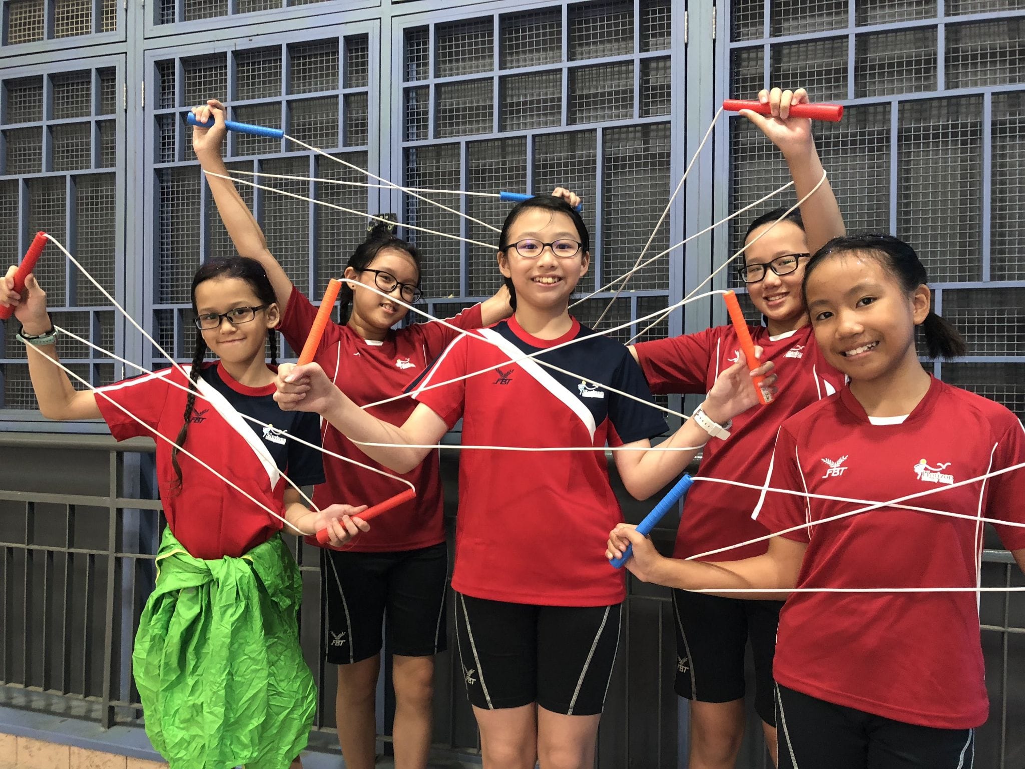 Five girls in red uniforms smile while holding colored handles connected by tangled white strings outdoors.