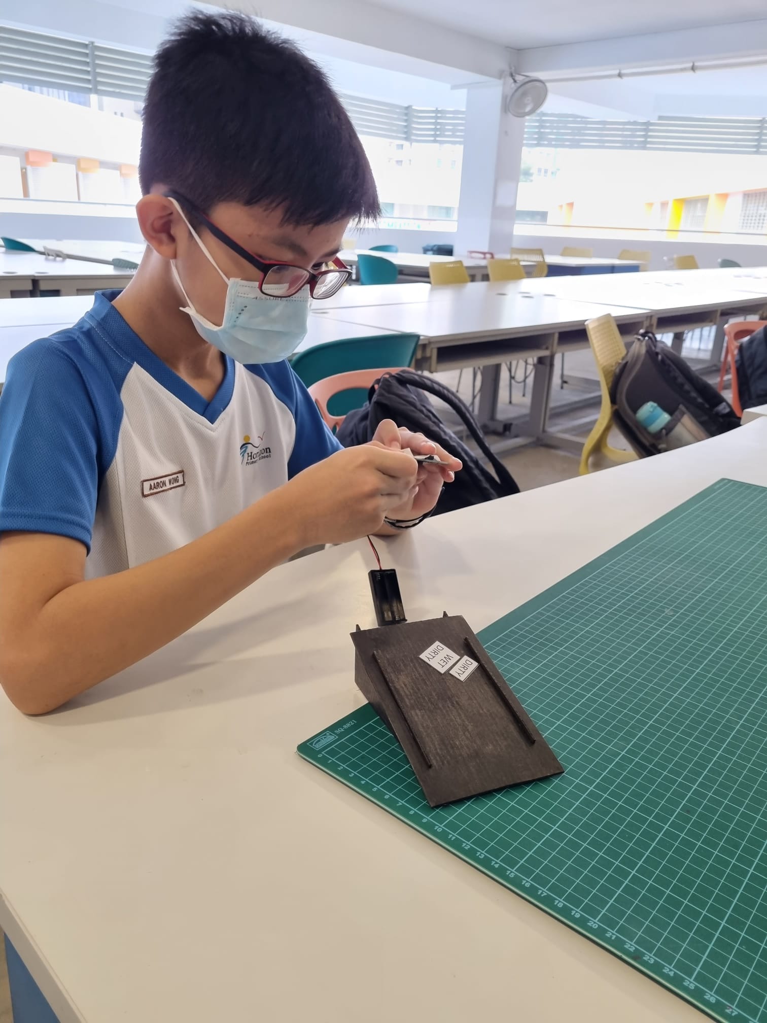 A masked boy wearing glasses works on a small wooden model with wires at a desk in a classroom.