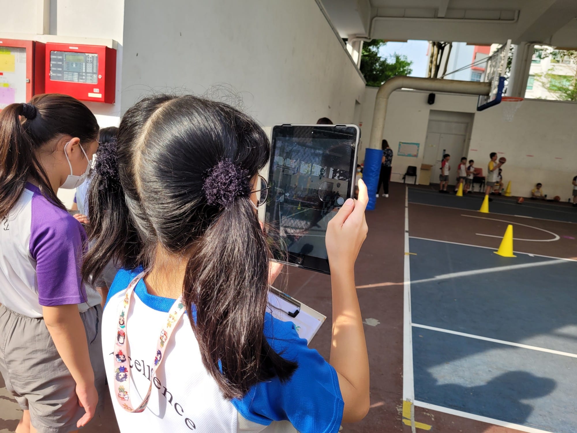 A girl holds up a tablet to photograph a school basketball court with yellow cones and students in the background.