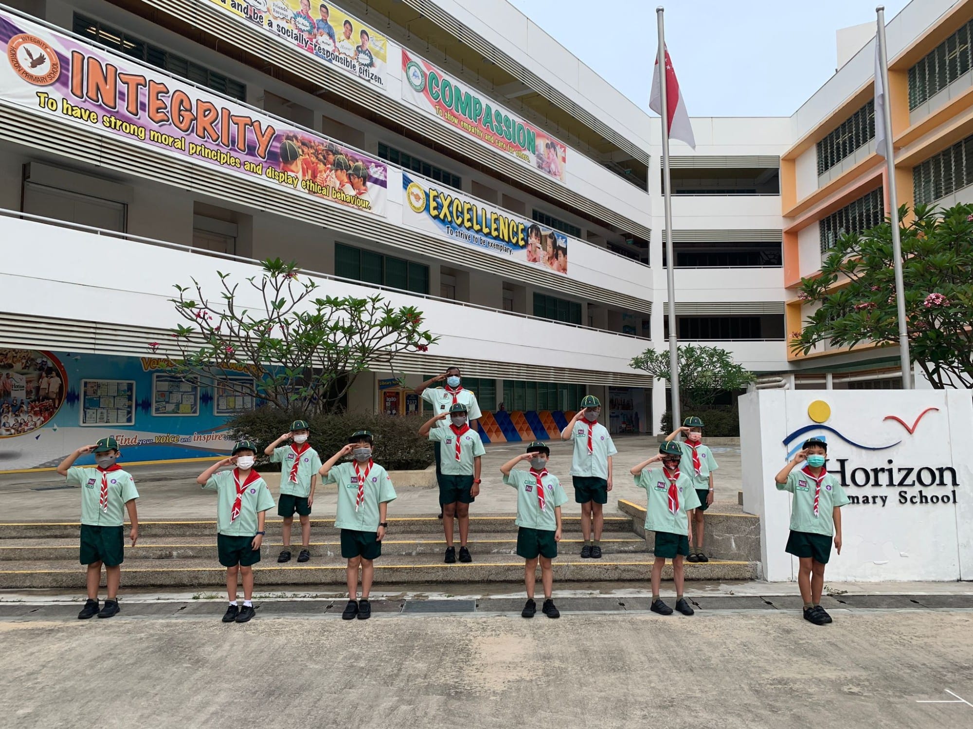 Young scouts in green uniforms and red scarves salute in front of Horizon Primary School in Singapore.