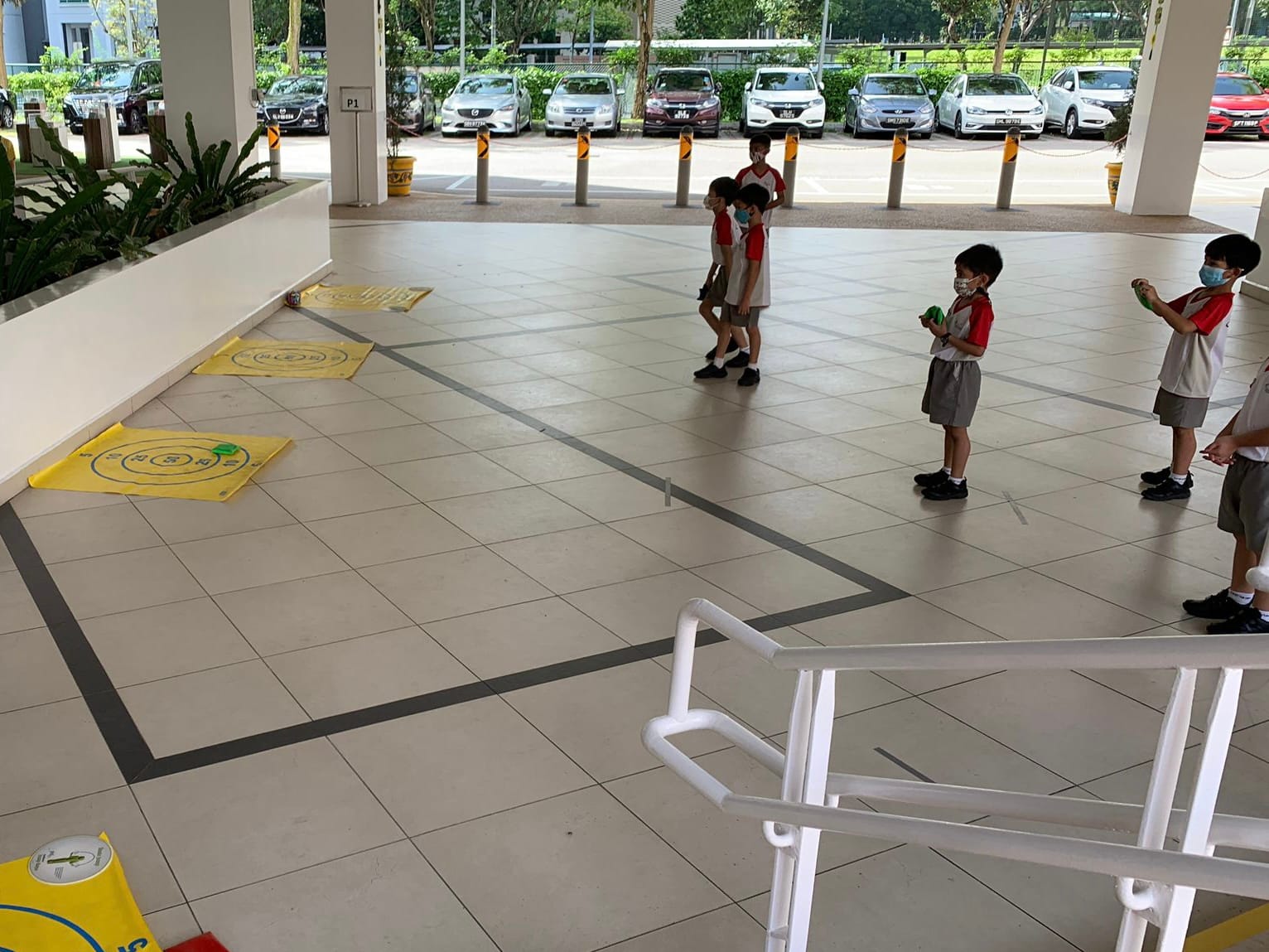 Children in school uniforms and masks play outdoors under a covered area with yellow target mats on the floor.