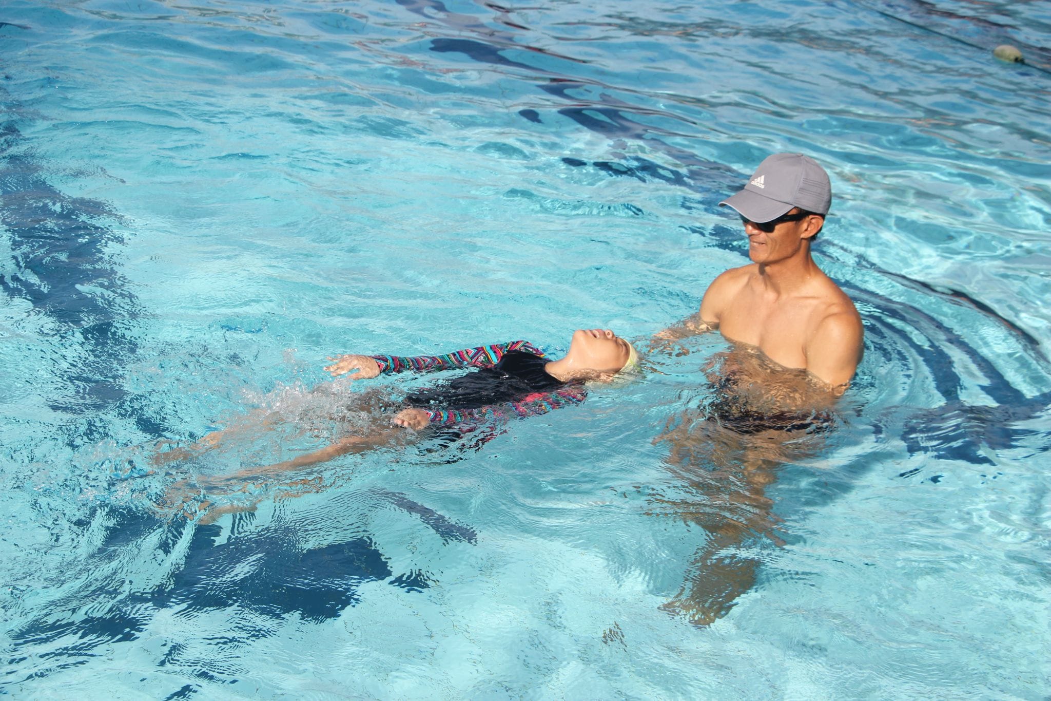 A man in a gray cap supports a child floating on their back during a swim lesson in a blue pool.