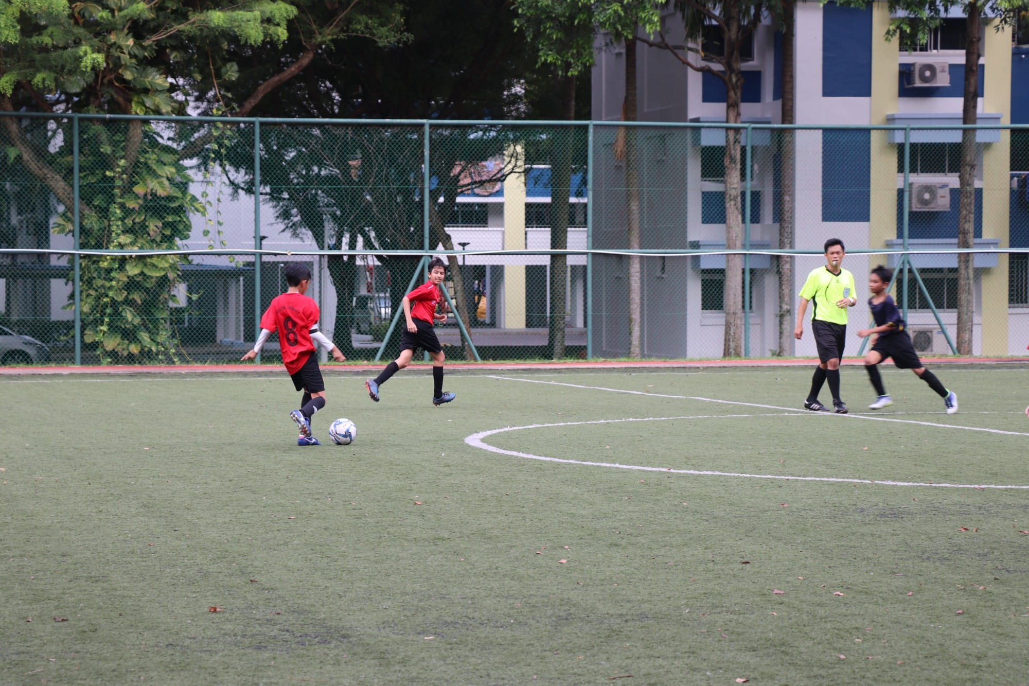 Young players in red jerseys compete on a green turf soccer field near a fenced court with buildings behind.