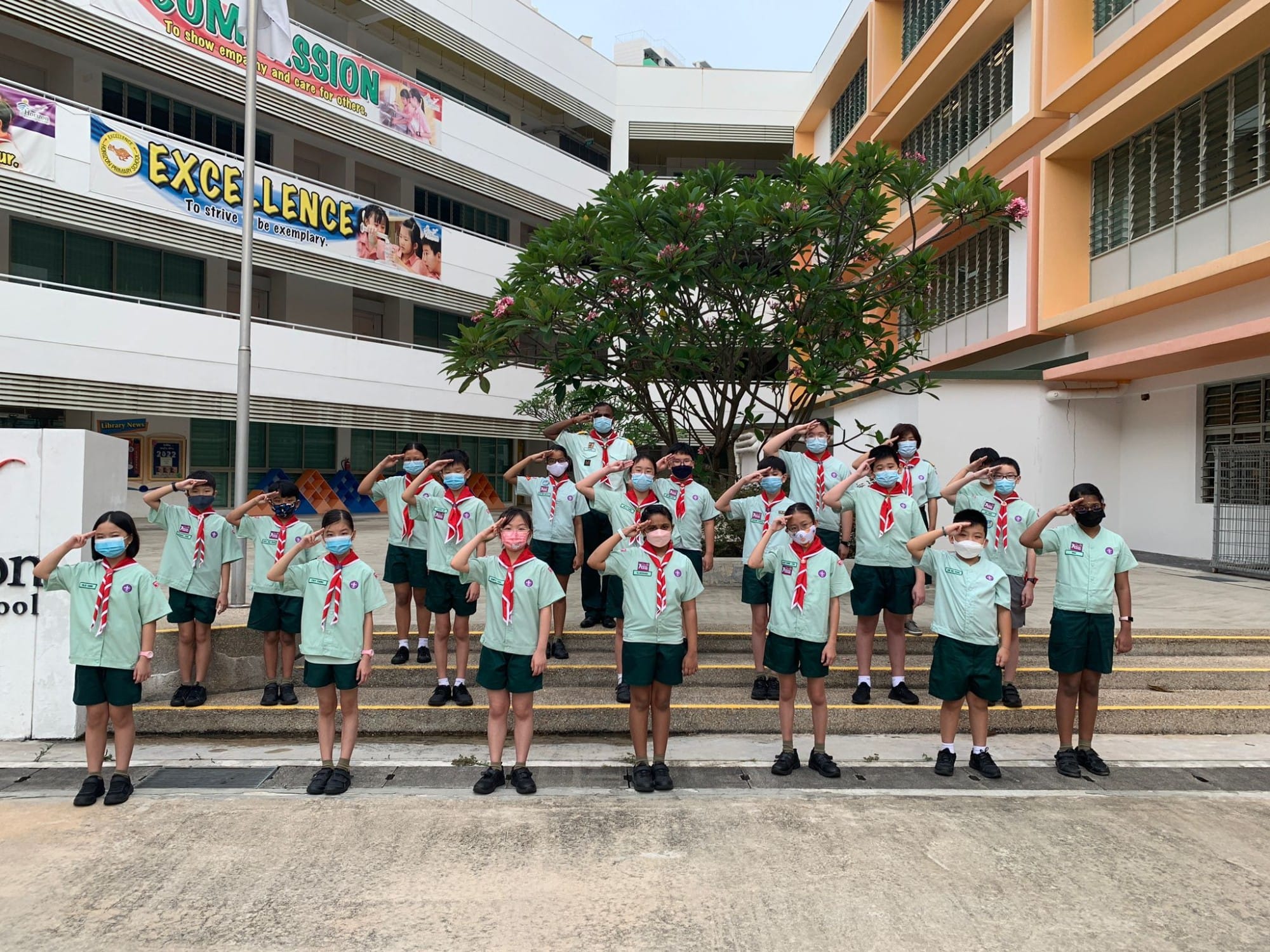 A group of children in green scout uniforms and red neckerchiefs saluting outdoors at a school courtyard.