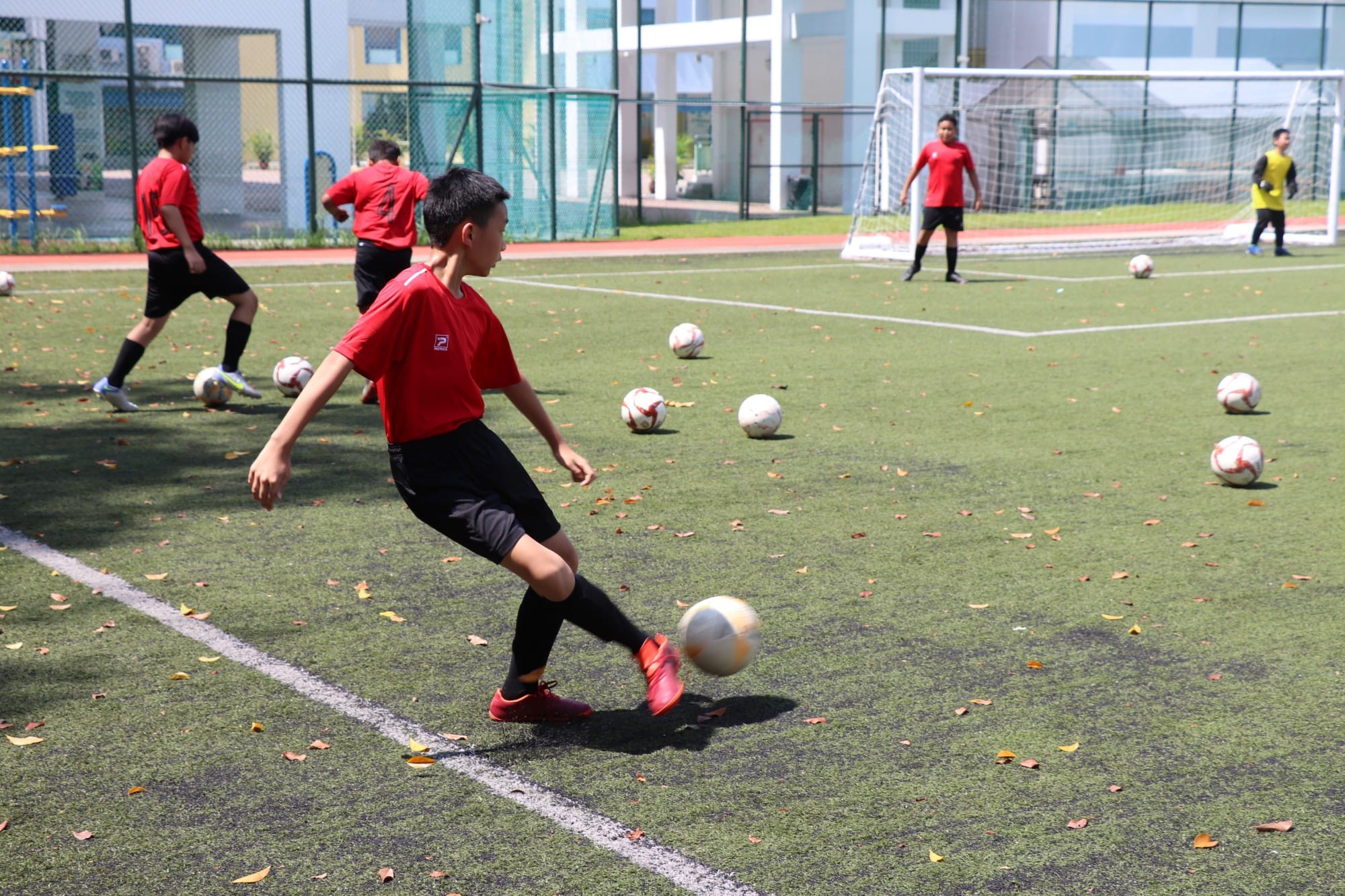 Boys in red uniforms practice kicking soccer balls on a green artificial turf field with a goal in the background.