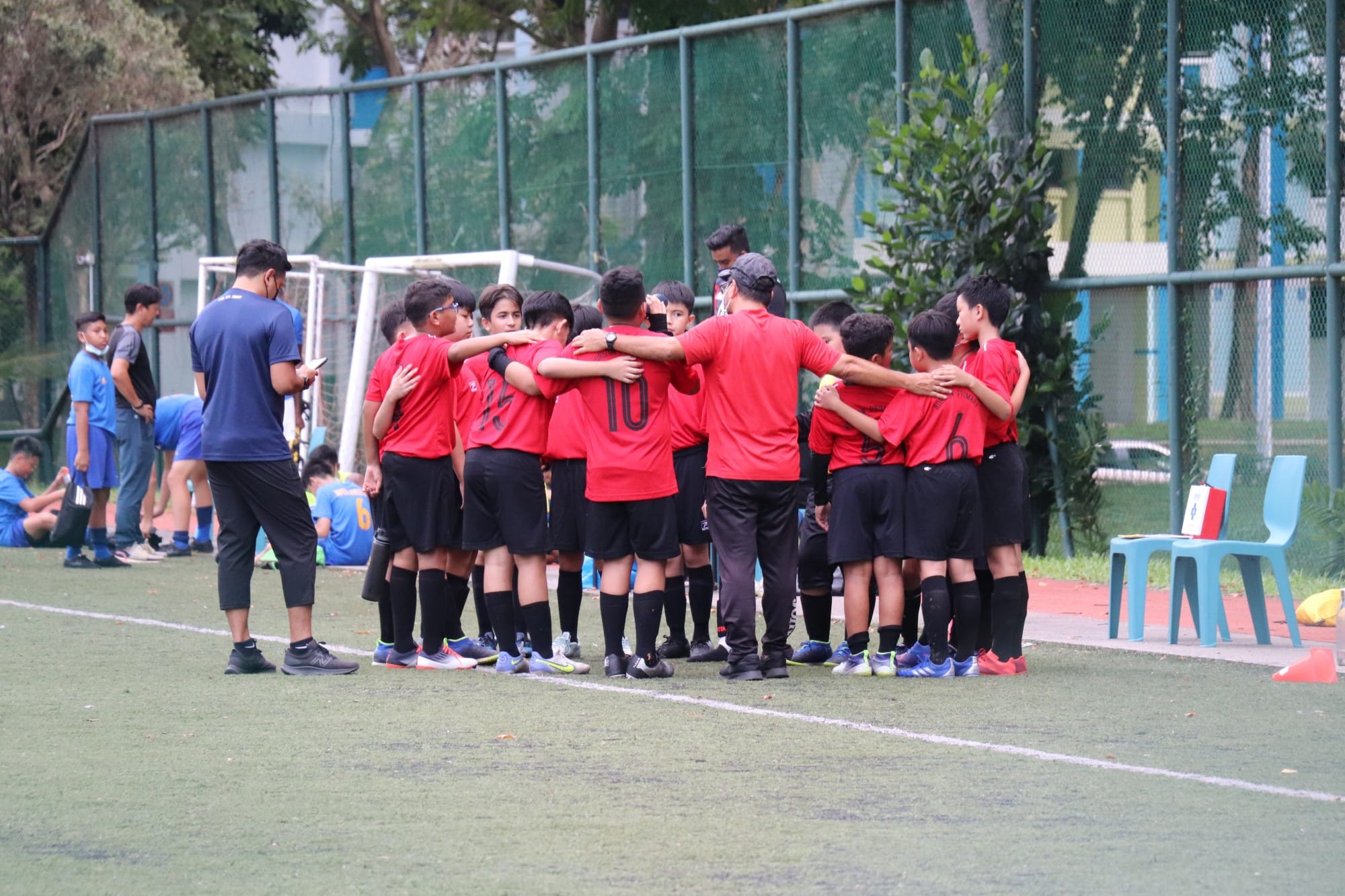 A youth soccer team in red and black uniforms huddles together with a coach on a green field near a goal.