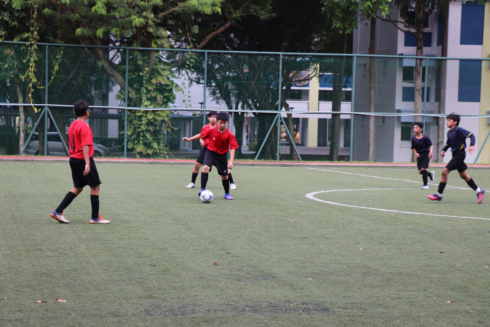 Five young boys play soccer on a green turf field, two in red jerseys and three in dark uniforms.
