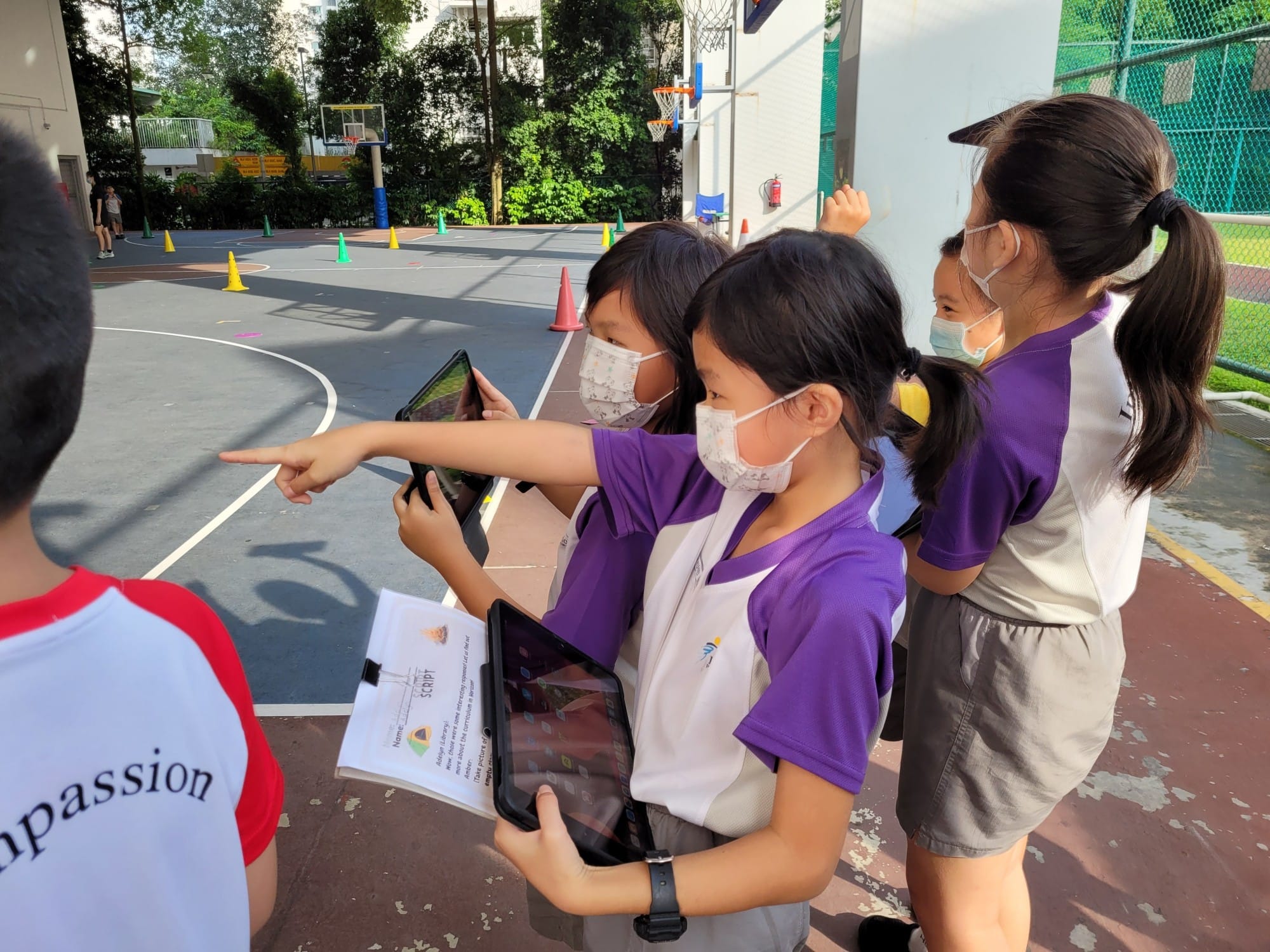 Children in purple uniforms and face masks hold tablets and point toward a basketball court with traffic cones.