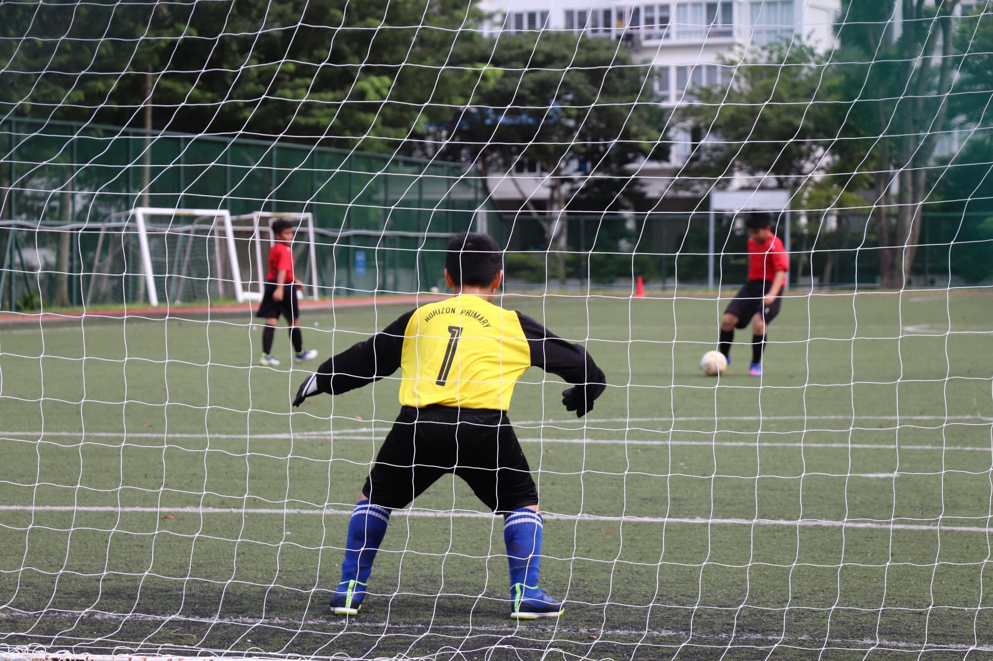 A young goalkeeper in yellow jersey number 1 stands ready in goal as a player in red prepares to shoot.