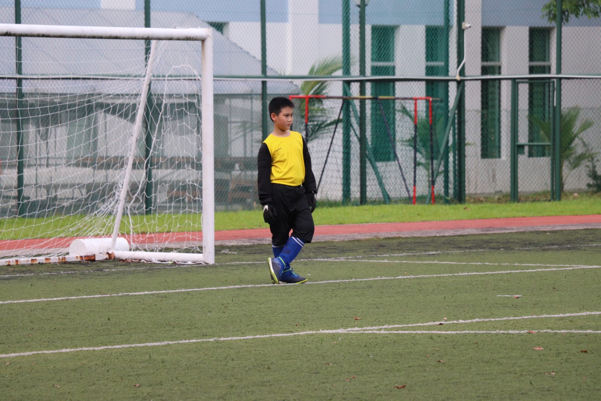 A young boy in a yellow vest and black goalkeeper gloves stands near a white football goal on a green pitch.