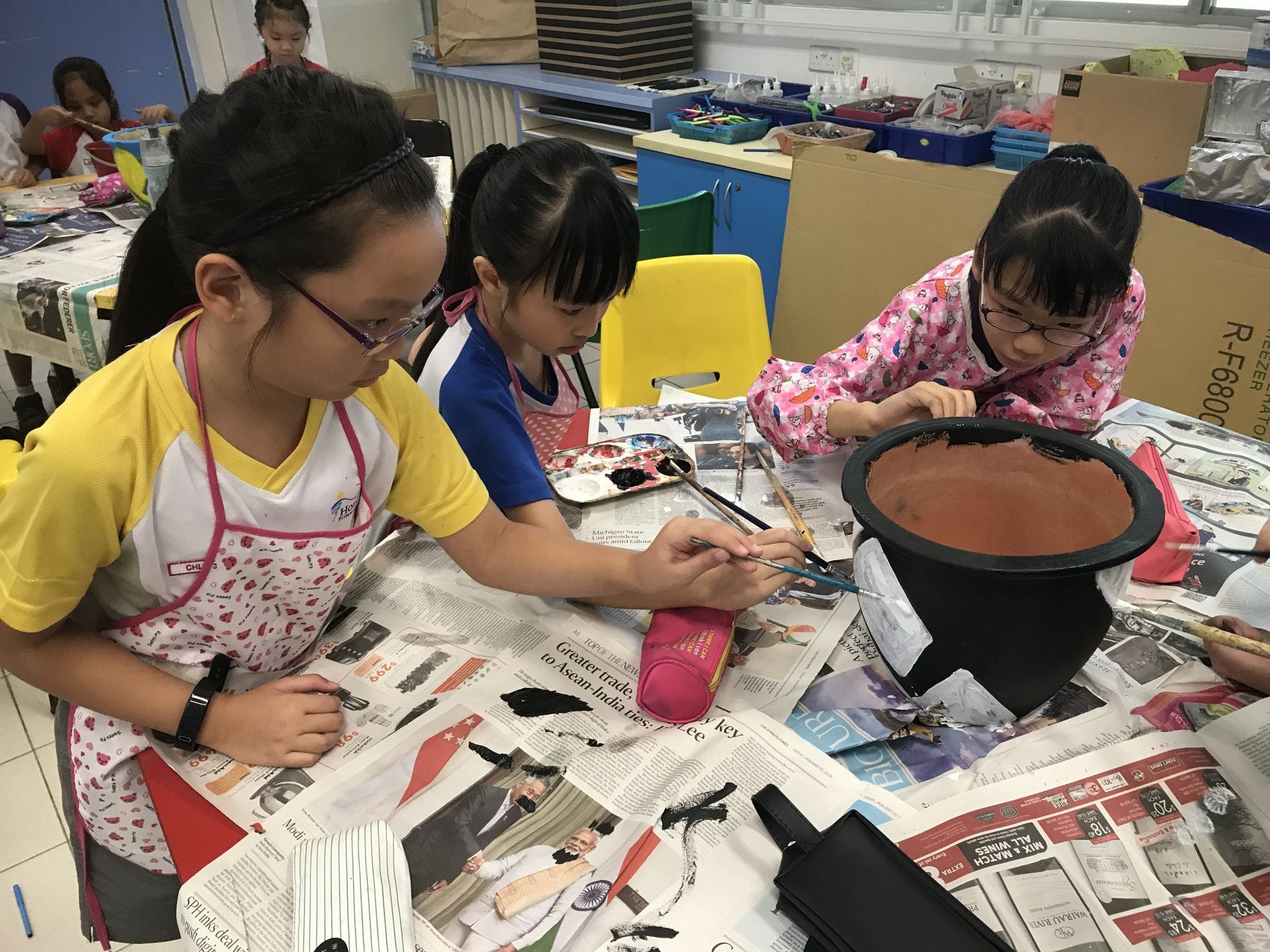 Three children in aprons paint a large black clay pot on a newspaper-covered table in an art classroom.