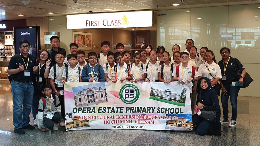 Group of students and teachers posing with an Opera Estate Primary School Vietnam cultural trip banner at an airport.