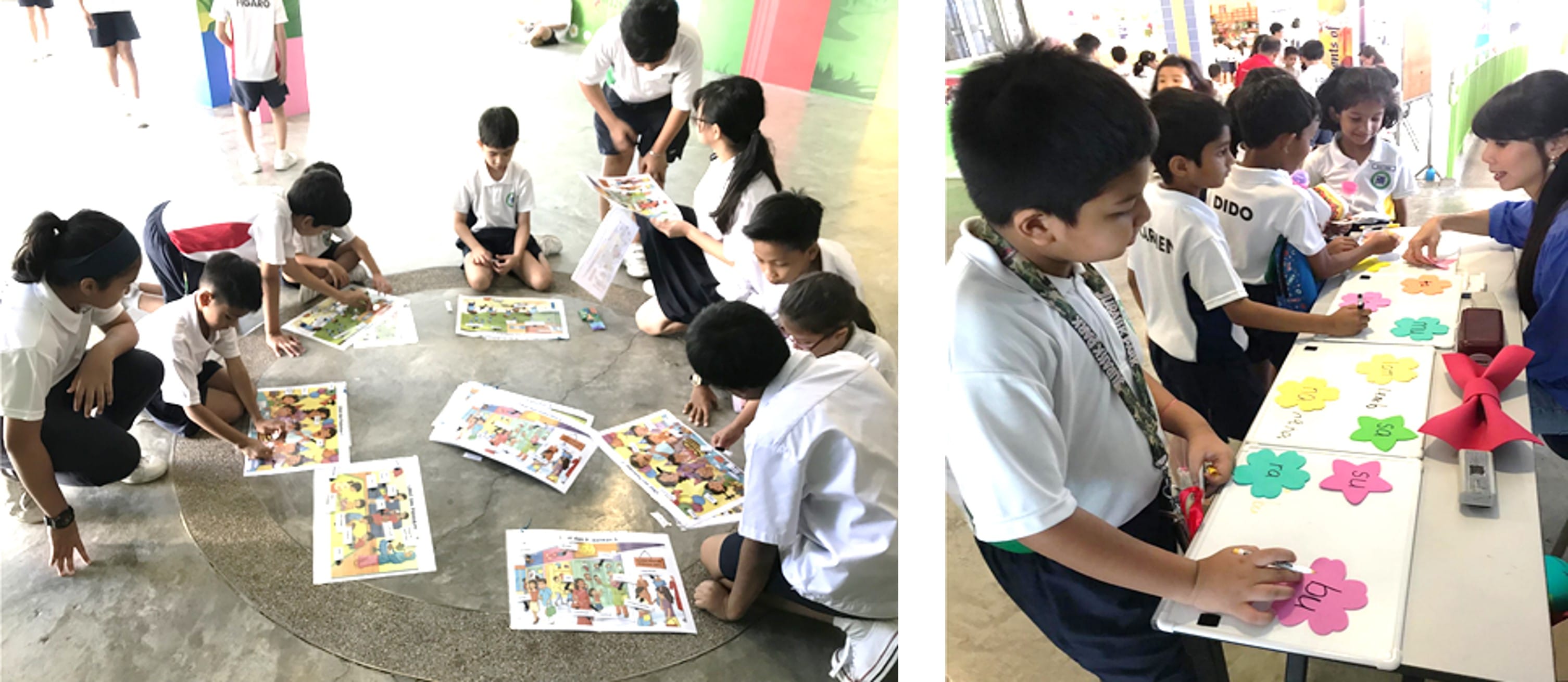 Children in uniforms sit in a circle examining colorful picture cards on the floor, while others write on flower-shaped paper cutouts.