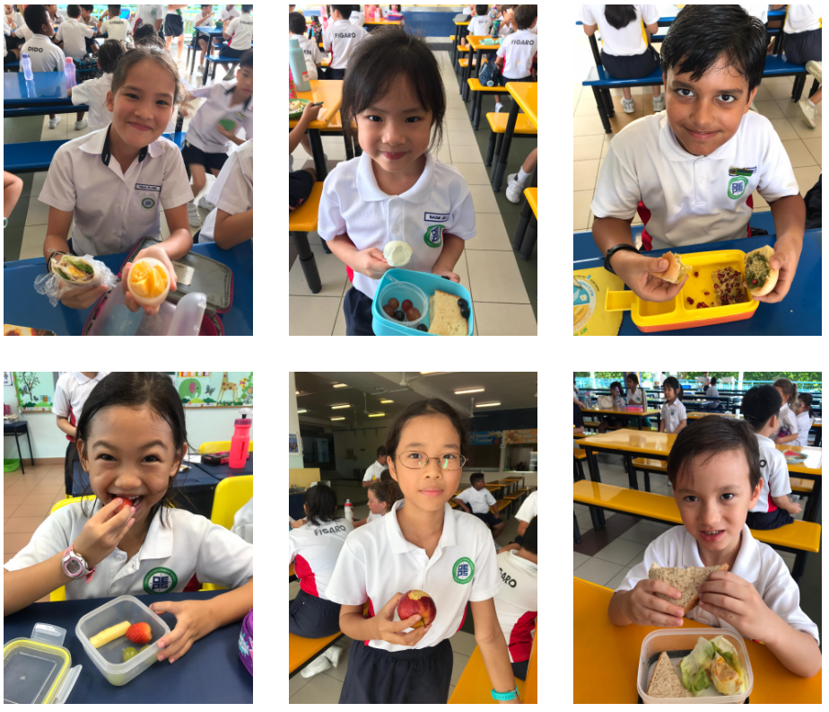 Six school children in uniforms smile while holding or eating lunch items like fruit, sandwiches, and lunchboxes in a cafeteria.