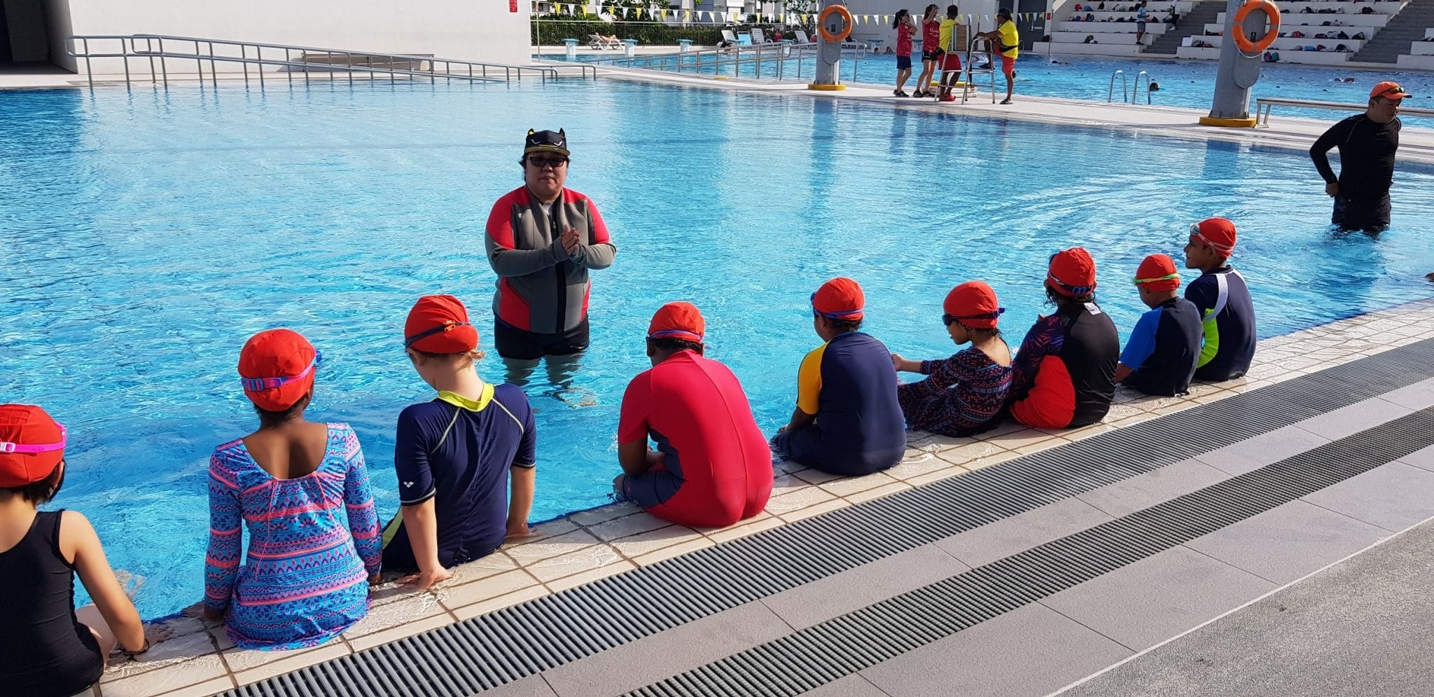 Children in red swim caps sit at the edge of a pool while an instructor stands in the water addressing them.