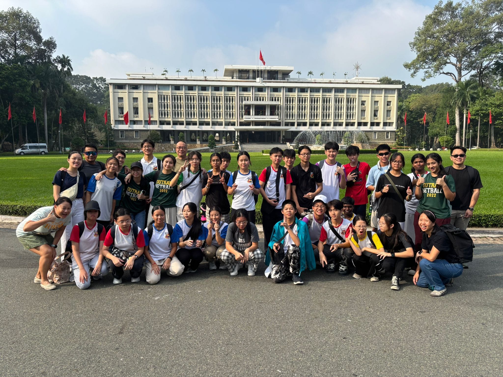 A large group of young people posing in front of a grand white building with red flags and a fountain.