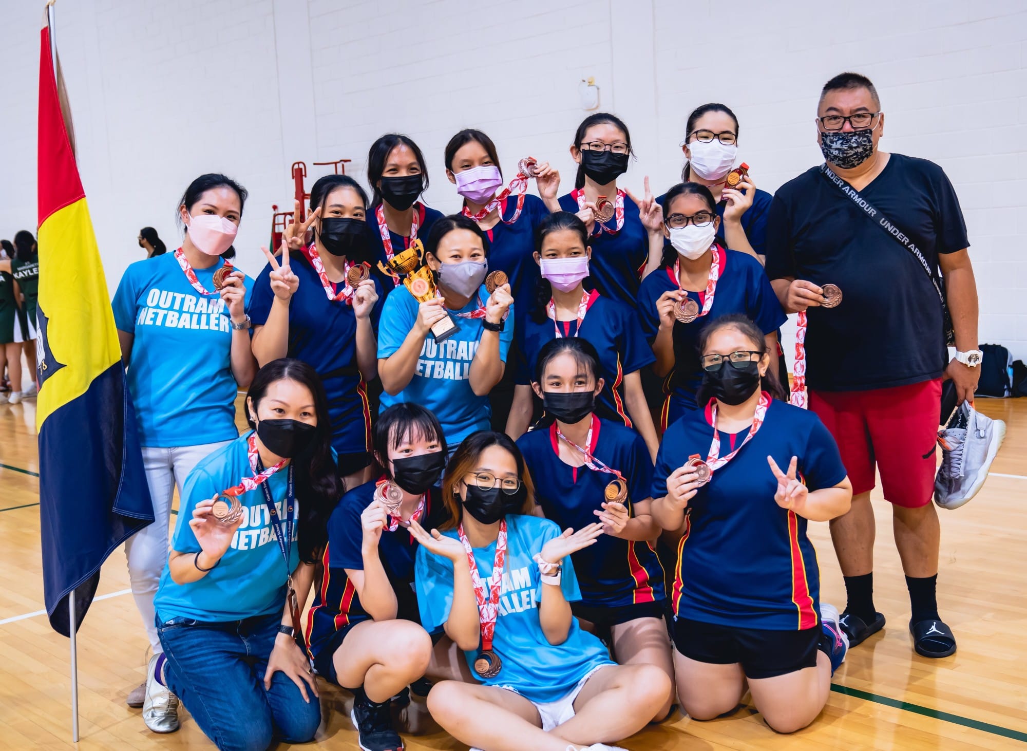 Outram Netballers team posing with medals and a trophy in a gym, all wearing face masks.