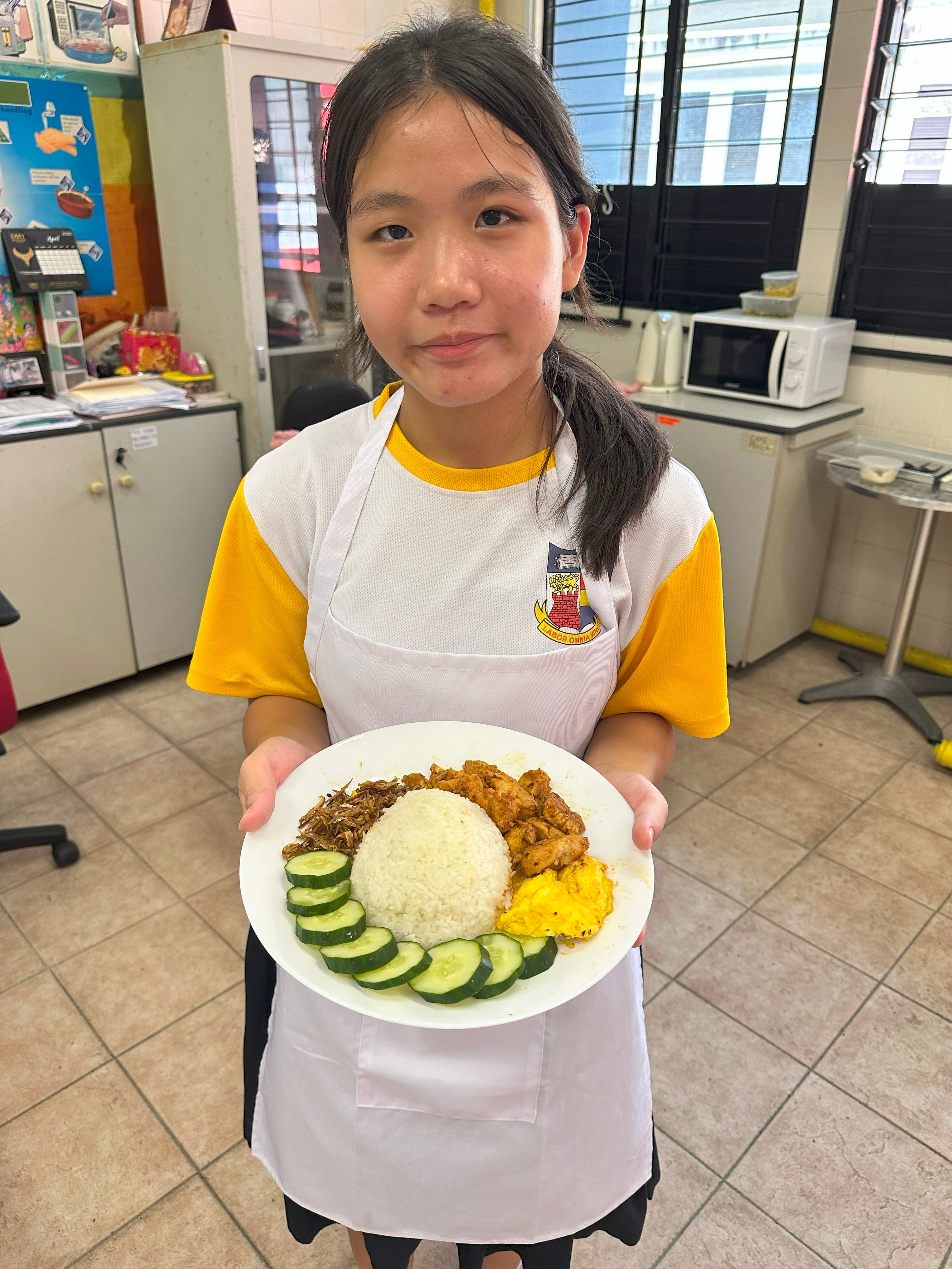 A young girl in a school uniform and apron holds a plate of nasi lemak with rice, cucumber, egg, and chicken.