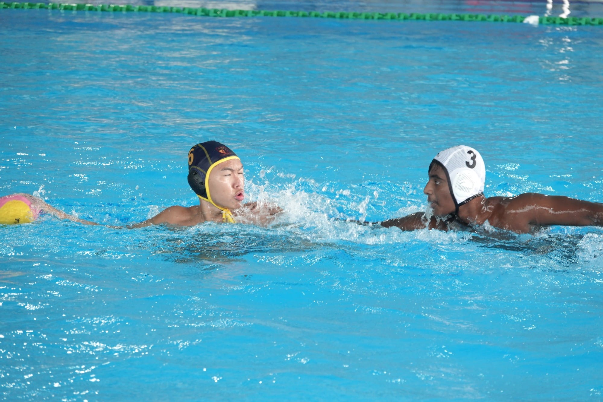 Two water polo players in caps numbered 6 and 3 compete near a yellow ball in a blue pool.