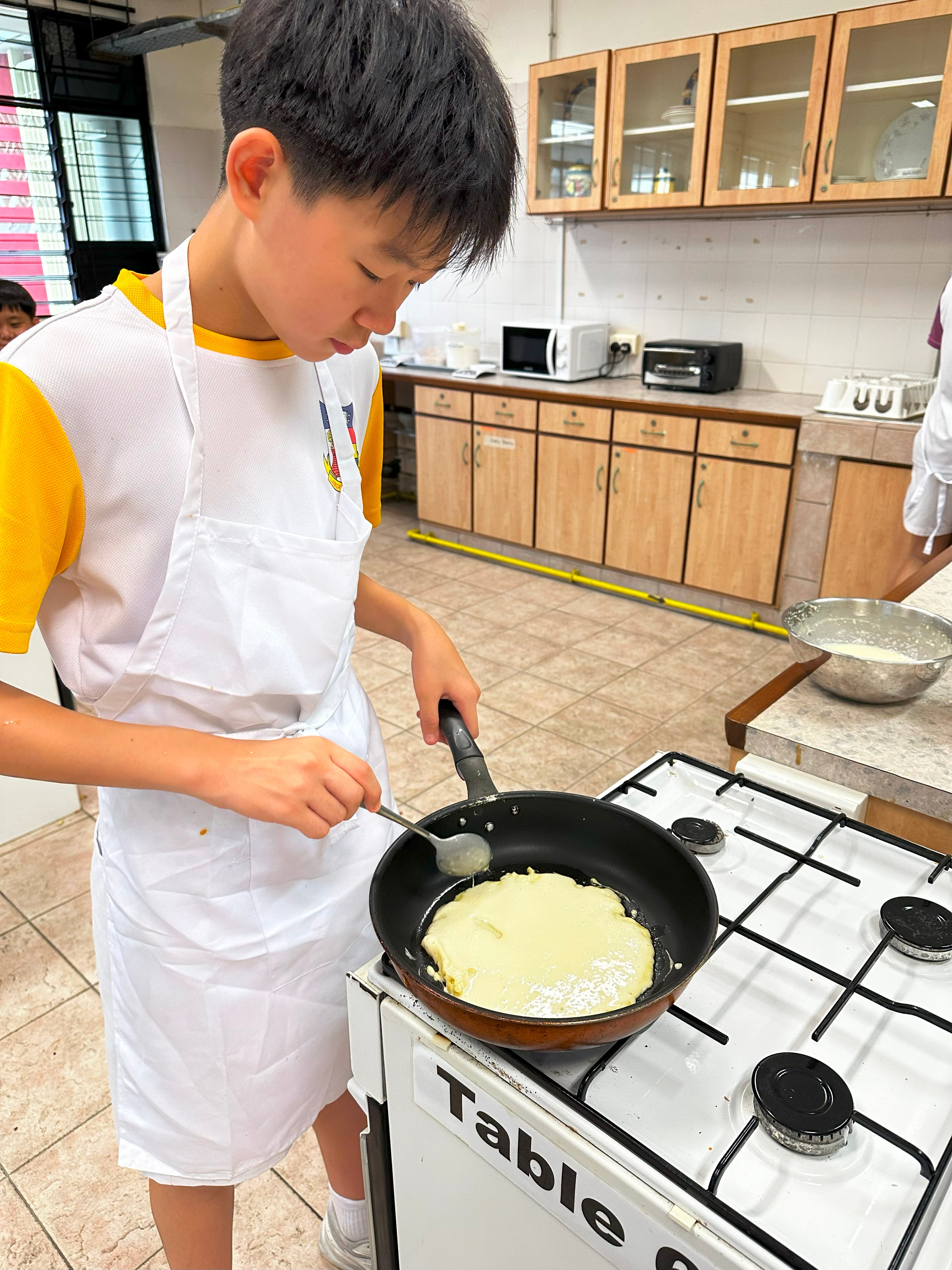 A boy in a white apron cooks a pancake in a black frying pan on a gas stove in a school kitchen.