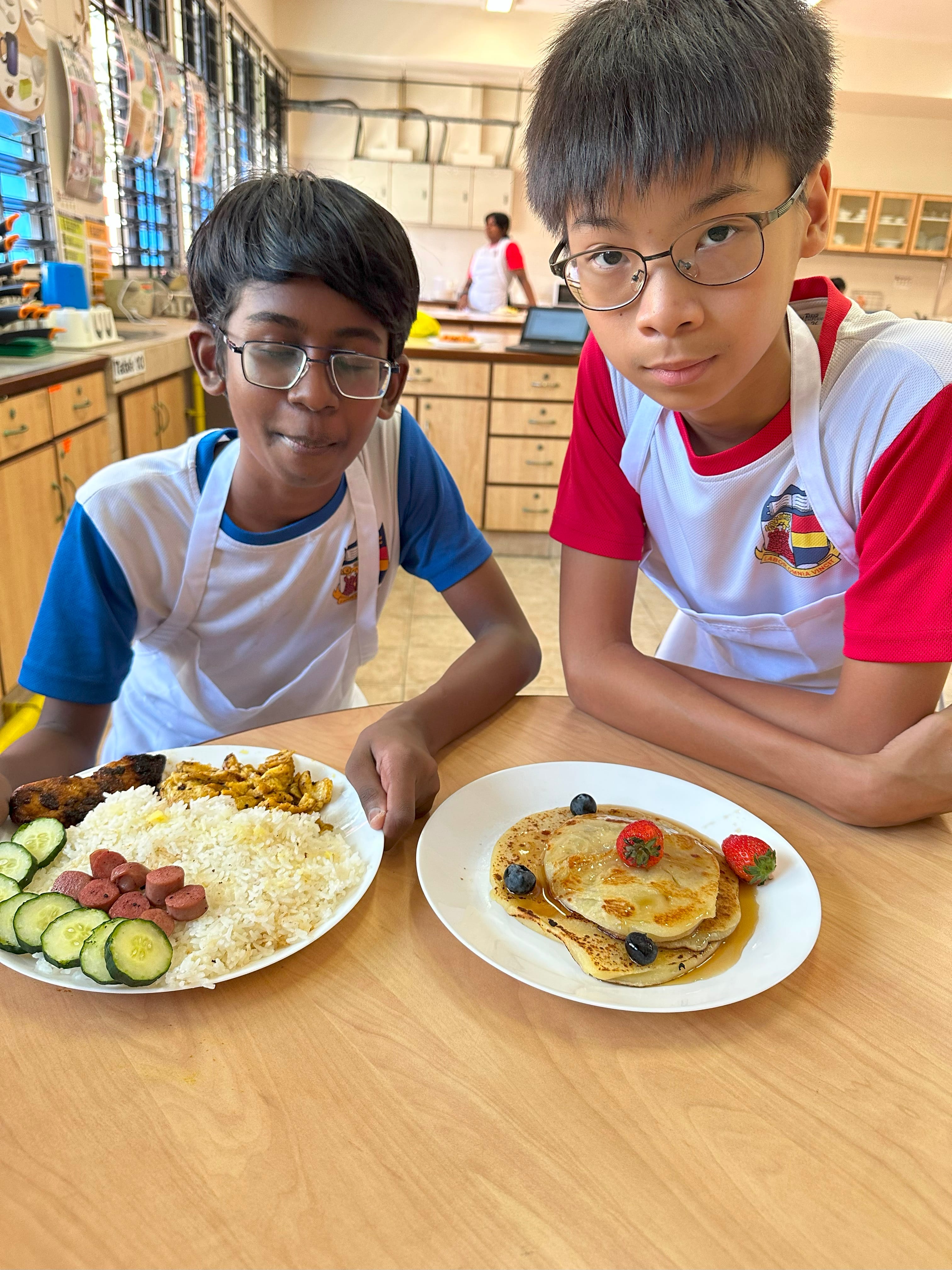 Two boys wearing aprons and glasses sit in a kitchen classroom with plates of rice with sides and pancakes with berries.
