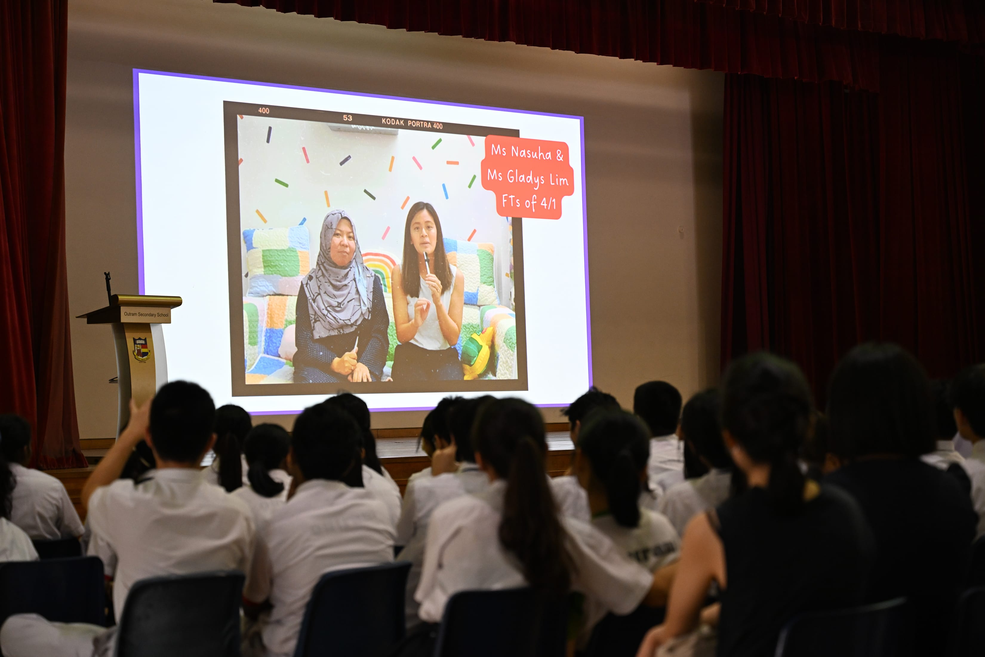 Students in white uniforms watch a projected slide showing two teachers, Ms Nasuha and Ms Gladys Lim, in a school hall.