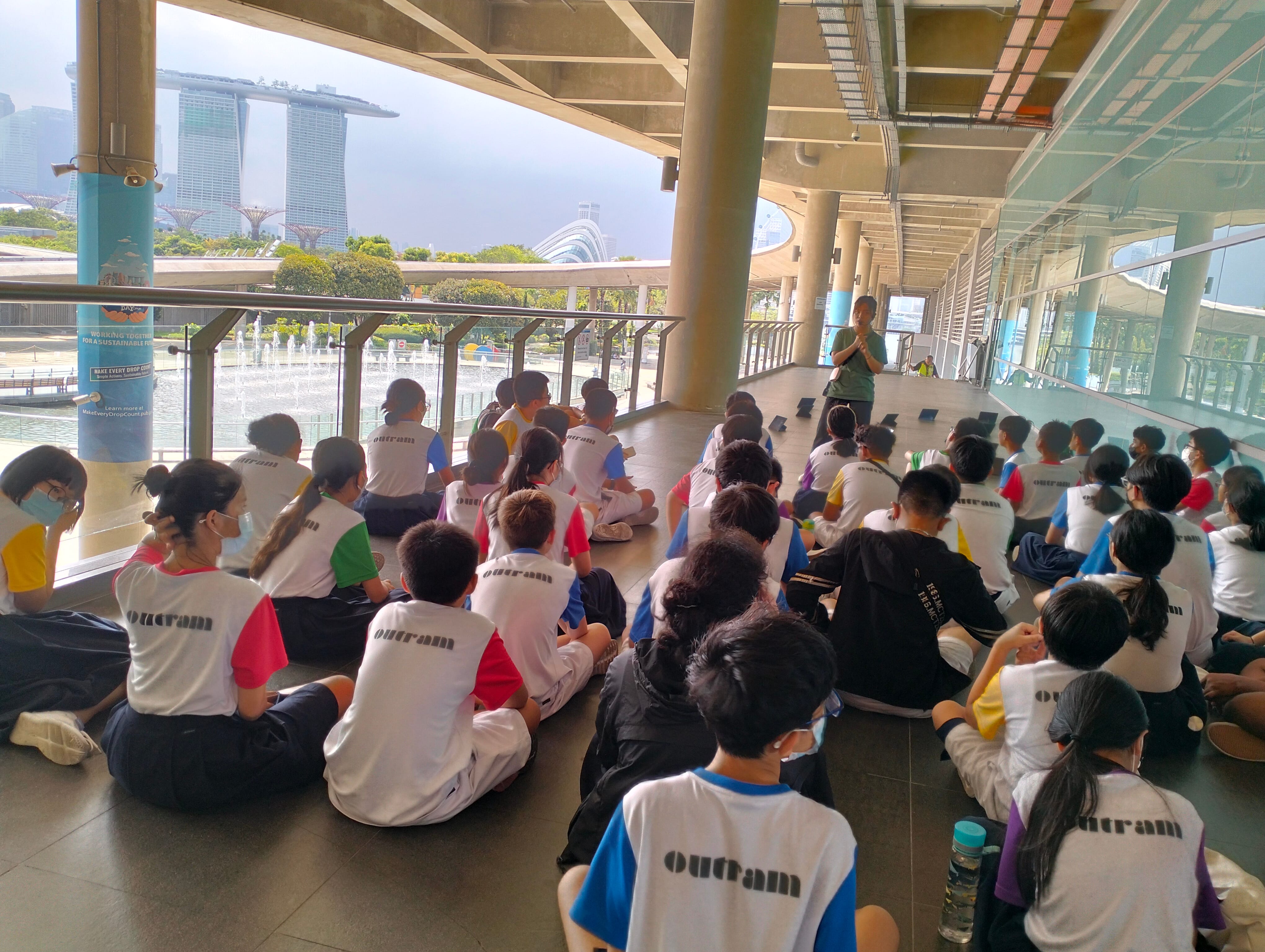 Students in multi-colored shirt sitting on the floor listening to a lady giving a briefing, at Marina Barrage.