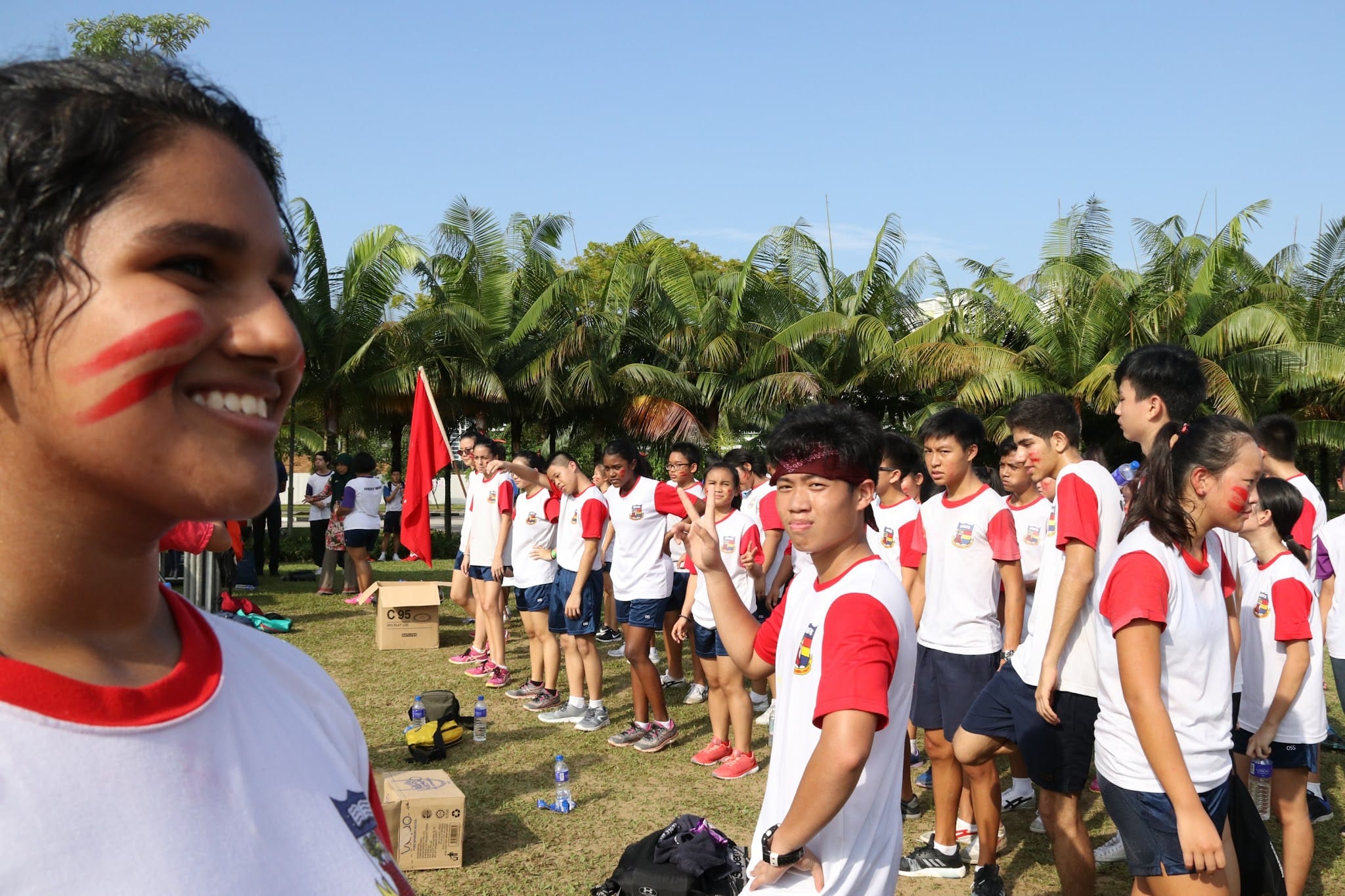 Students in white and red uniforms with red face paint stand on a grass field near palm trees during an outdoor event.