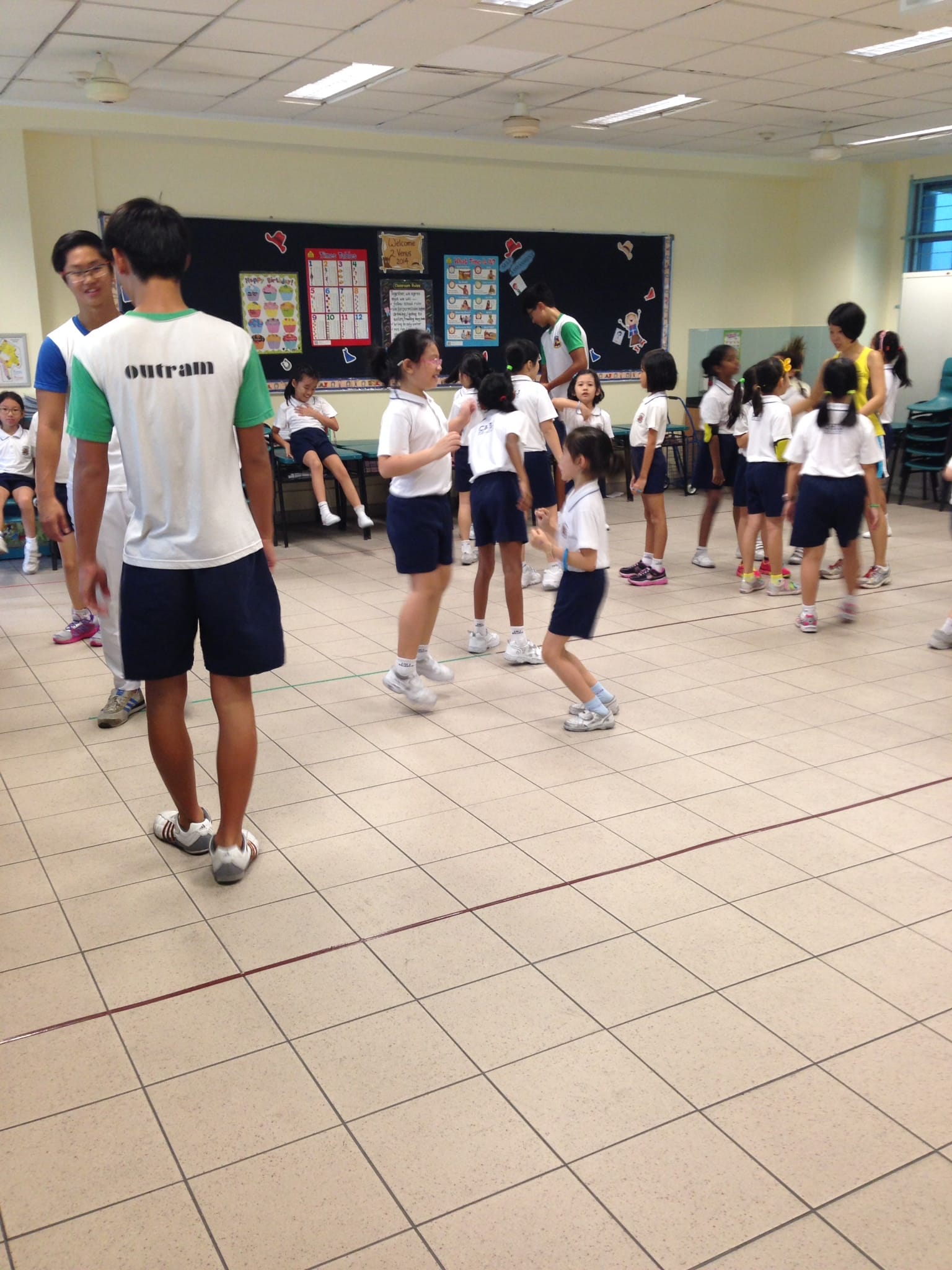 Children in school uniforms are active in an indoor hall, supervised by older students wearing Outram jerseys.