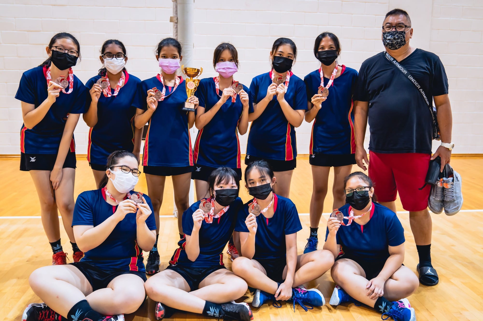 A girls sports team in navy uniforms and face masks pose with medals and a trophy on an indoor court.
