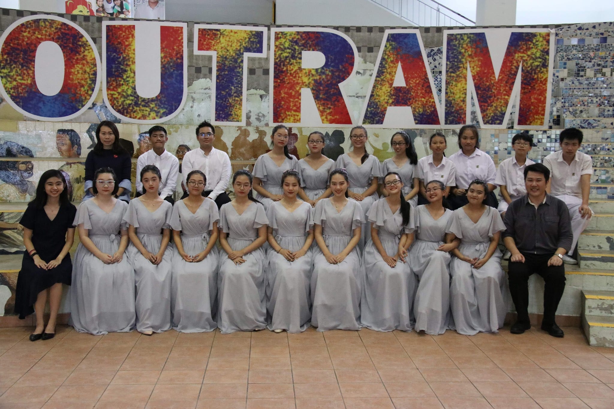 Group of young people posing in front of a colorful OUTRAM sign, most girls wearing matching silver-gray dresses.
