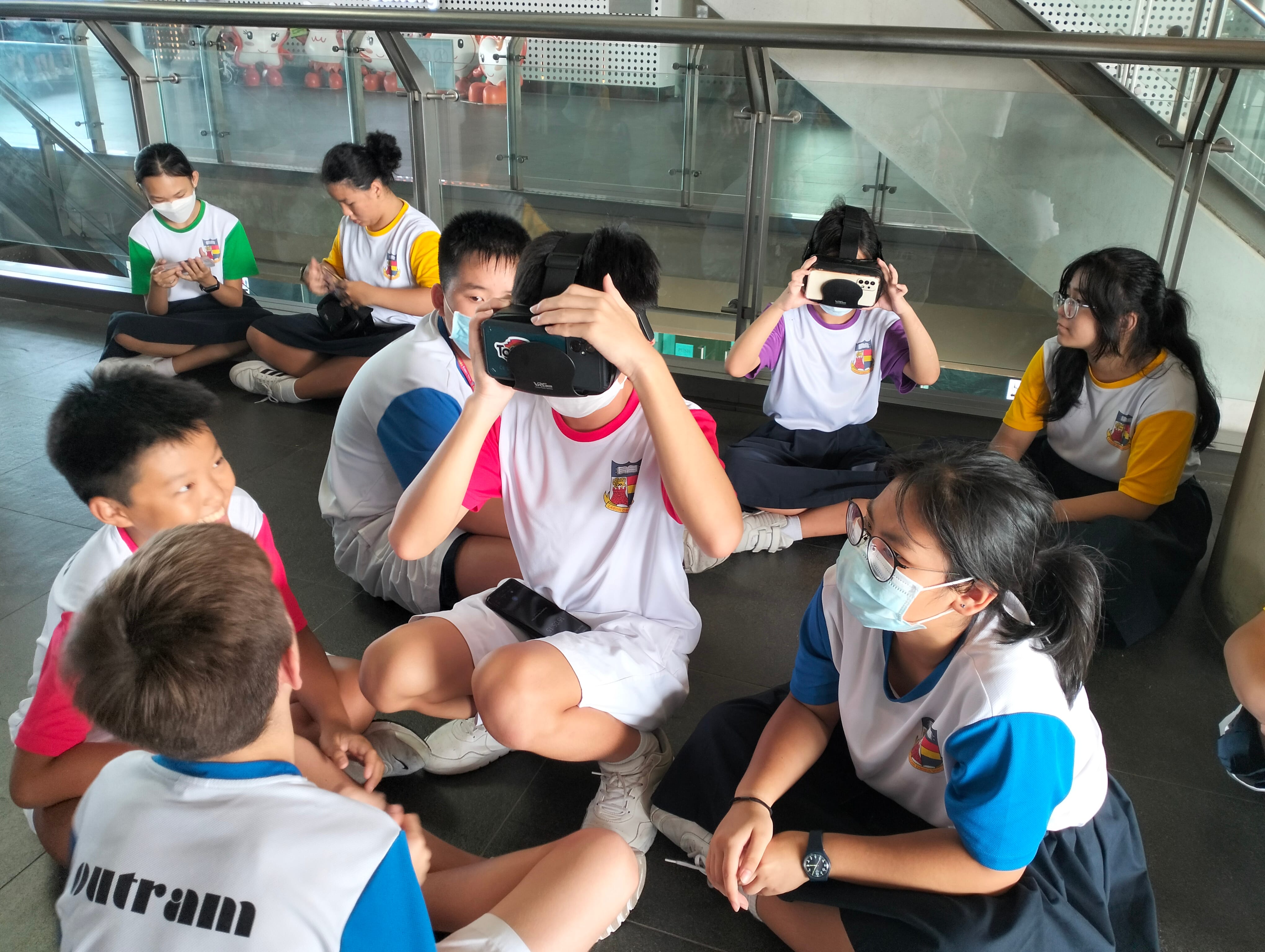 Students in school uniforms sit on a floor, some using VR headsets, others watching or waiting nearby.