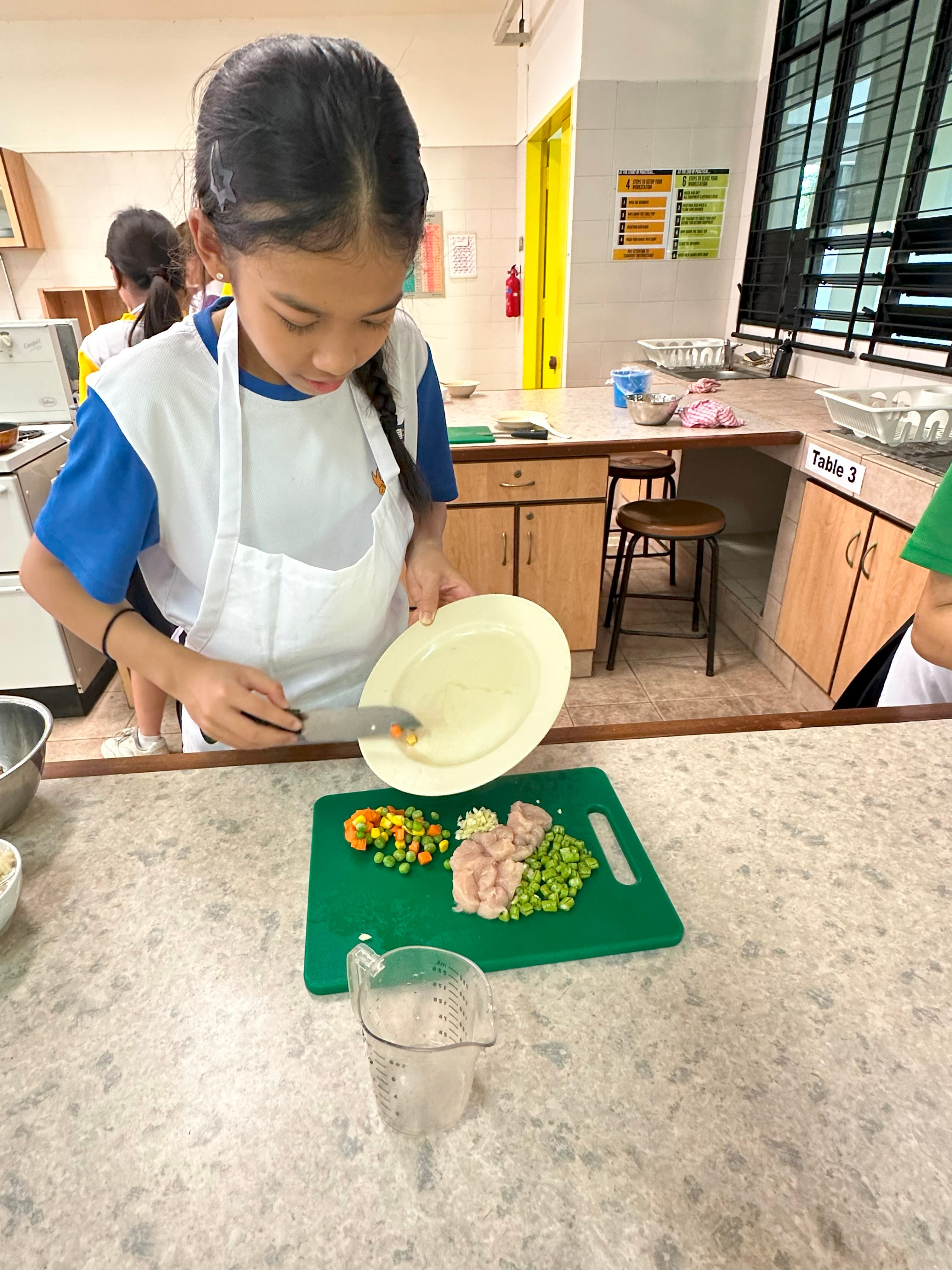 A young girl in a white apron tips mixed vegetables from a plate onto a green cutting board with meat and peas.