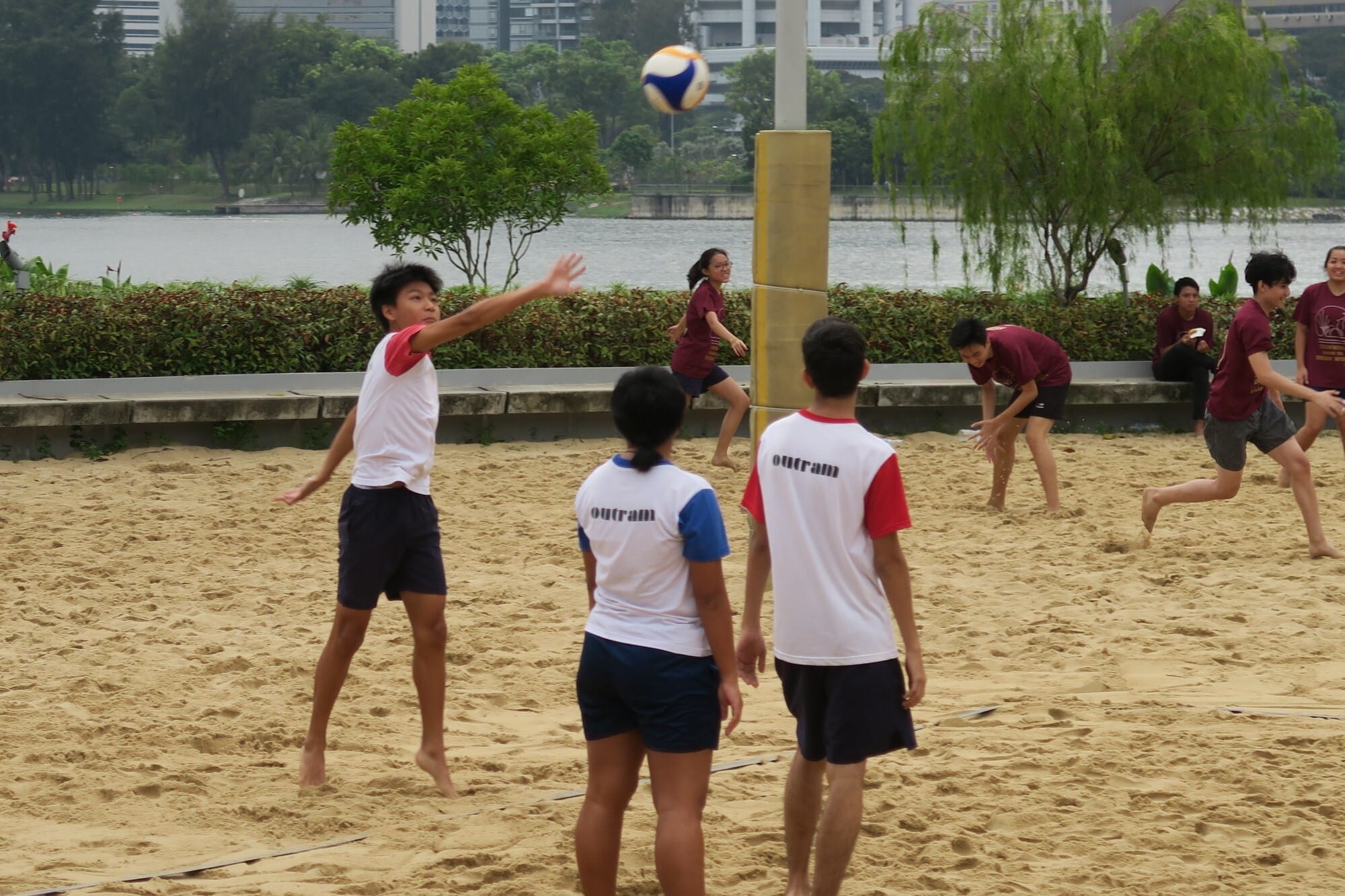 Youth players in Outram jerseys playing beach volleyball on sand, with a river and trees in the background.
