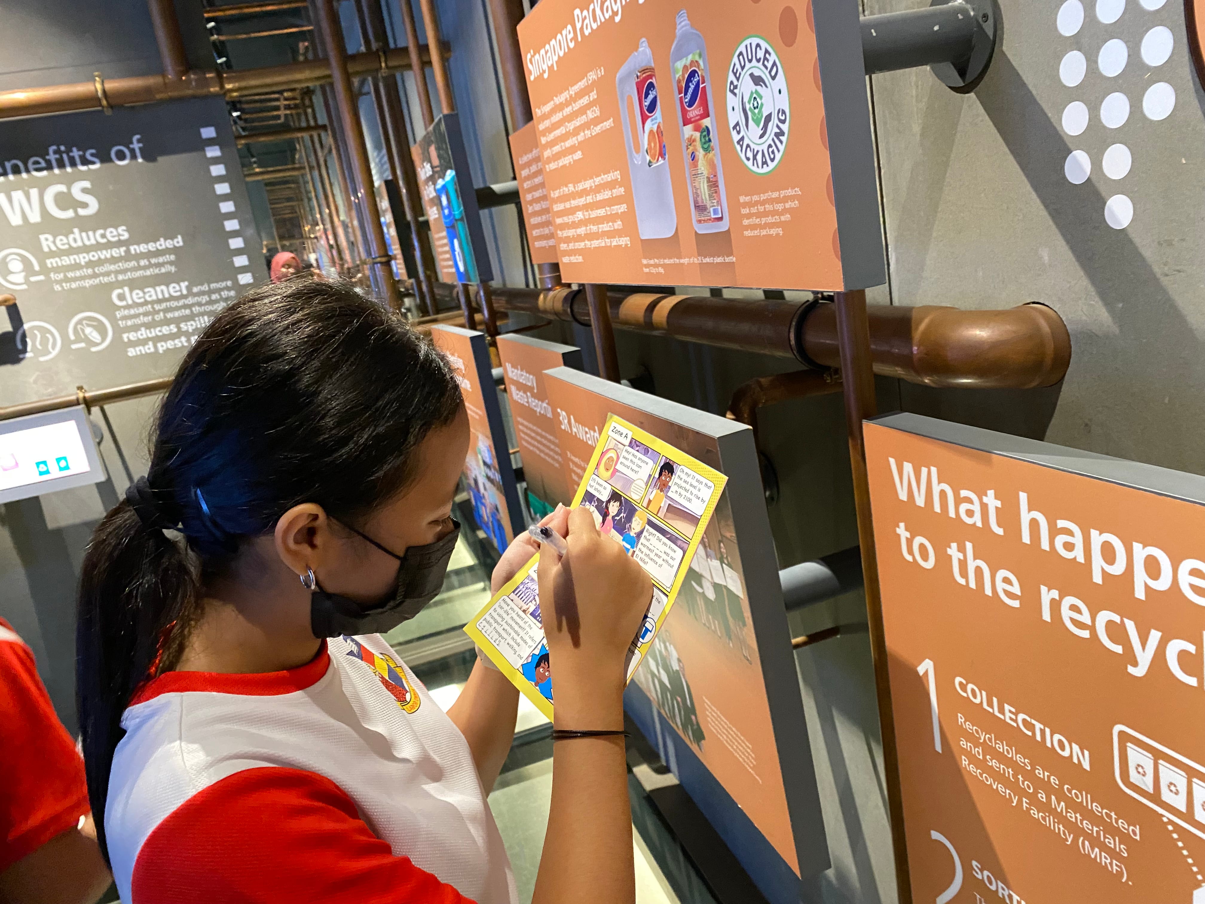 A masked girl writes on a comic booklet at a recycling museum exhibit with orange informational signs.