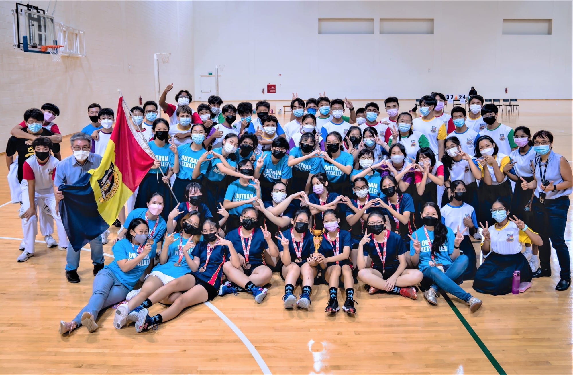 Large group of masked students in blue and white shirts pose with a trophy and flag on an indoor gym court.