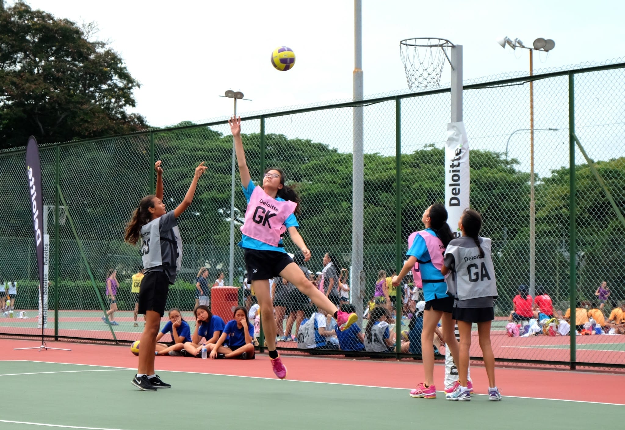 Girls playing netball outdoors, with one player in a pink GK bib jumping to intercept a ball near the hoop.