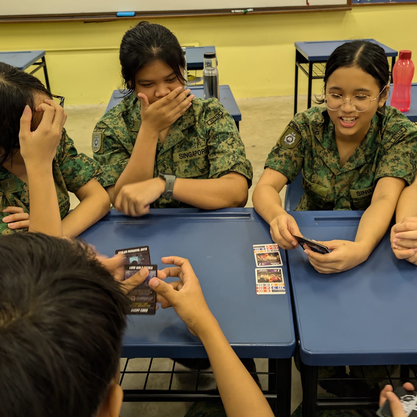 Cadets in camouflage uniform sit at desks playing a card game, reacting with laughter and surprise as they look at their cards.