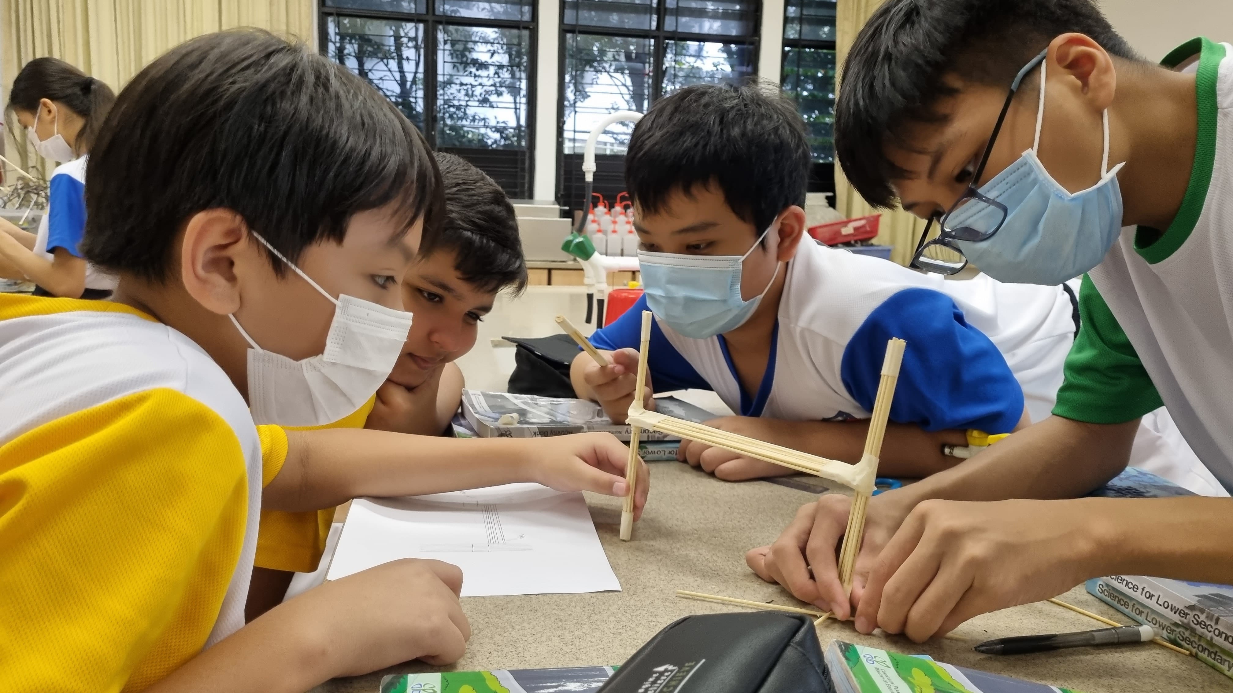 Four masked students lean over a desk, assembling a small wooden stick structure during a classroom activity.