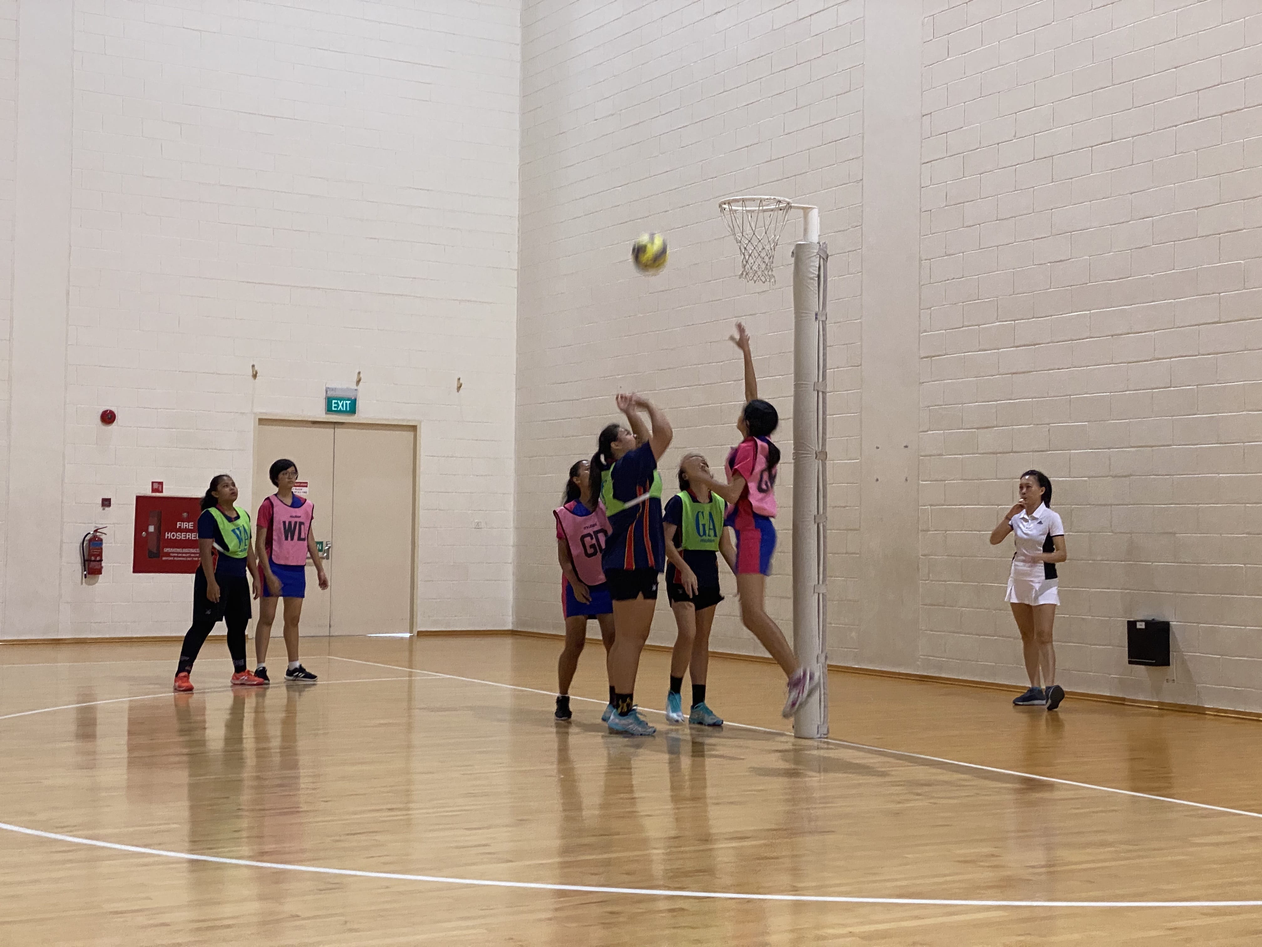Girls in pink and navy bibs play indoor netball, with one player jumping to shoot at the hoop.